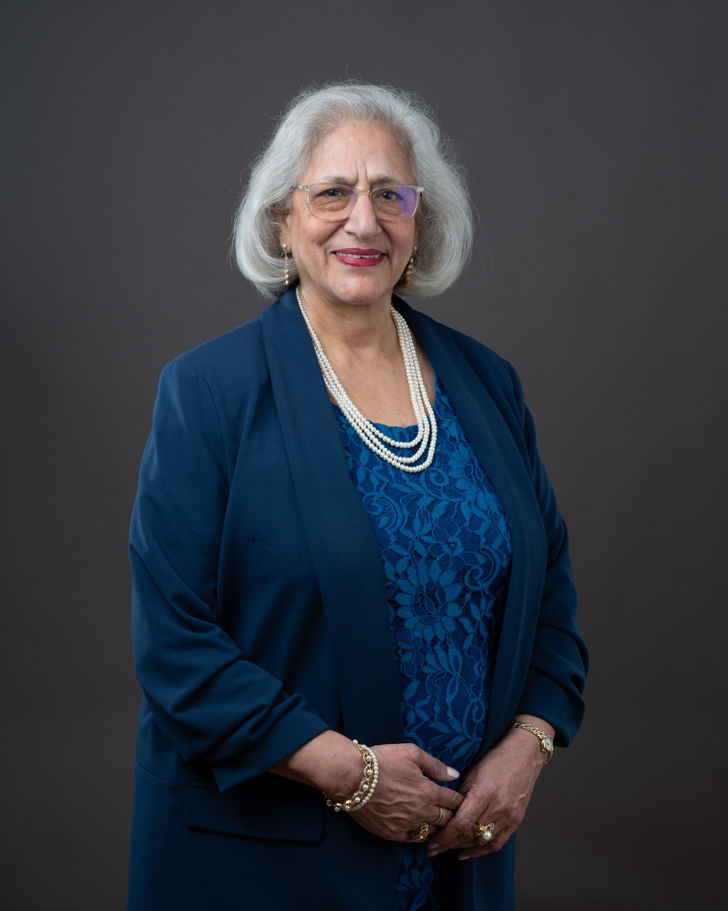 A person in a navy blue blazer and coordinating blue top poses for a professional portrait against a dark background.