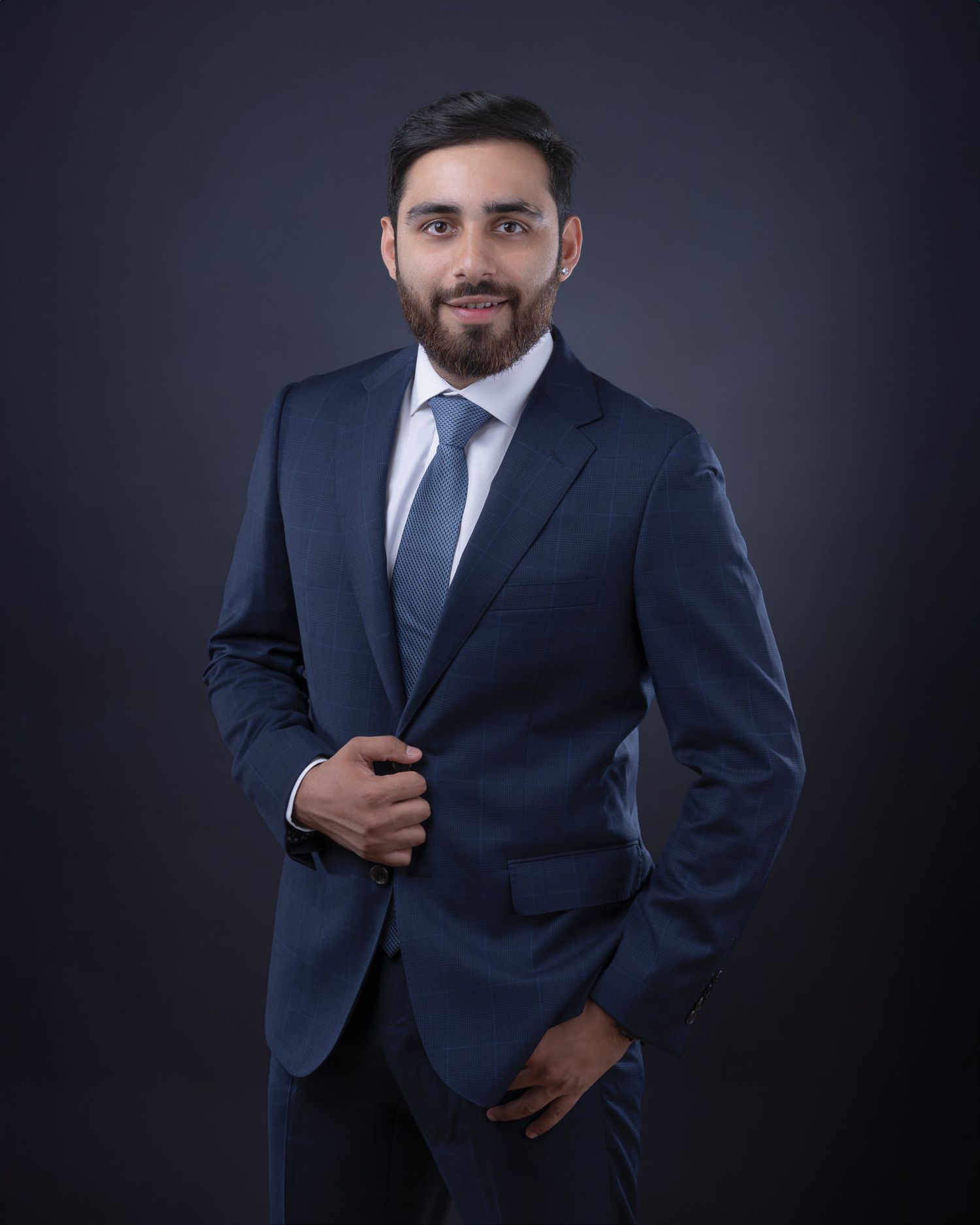 Professional business portrait of person in navy suit and tie against dark background, smiling confidently.