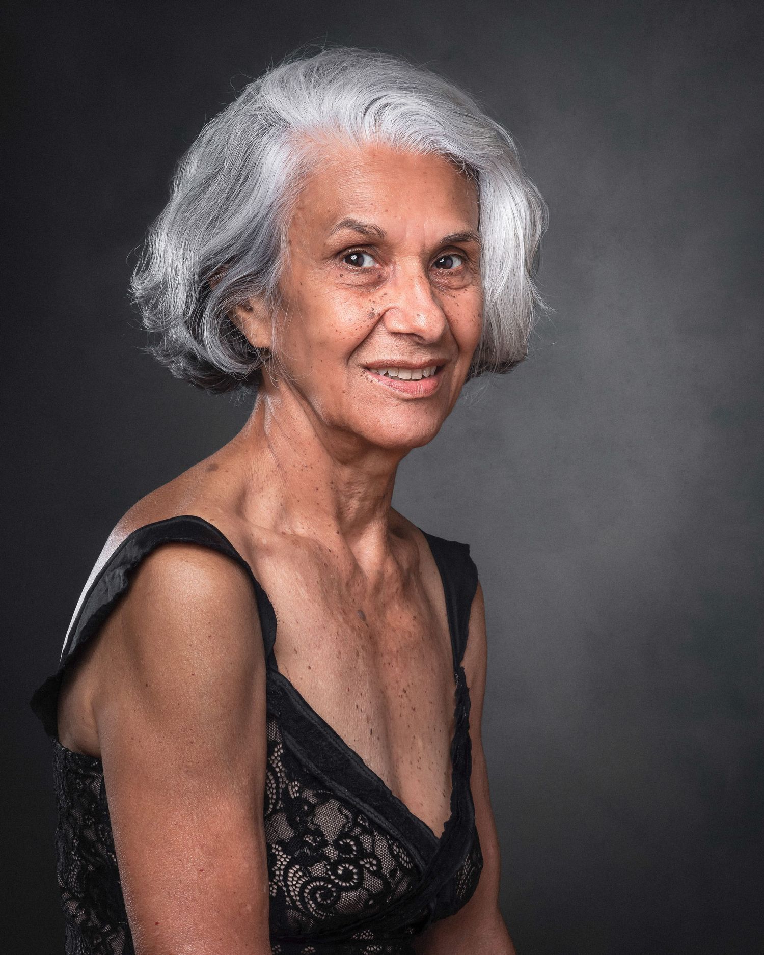 Elegant senior woman with silver hair wearing a black dress poses confidently against dark background in studio portrait.