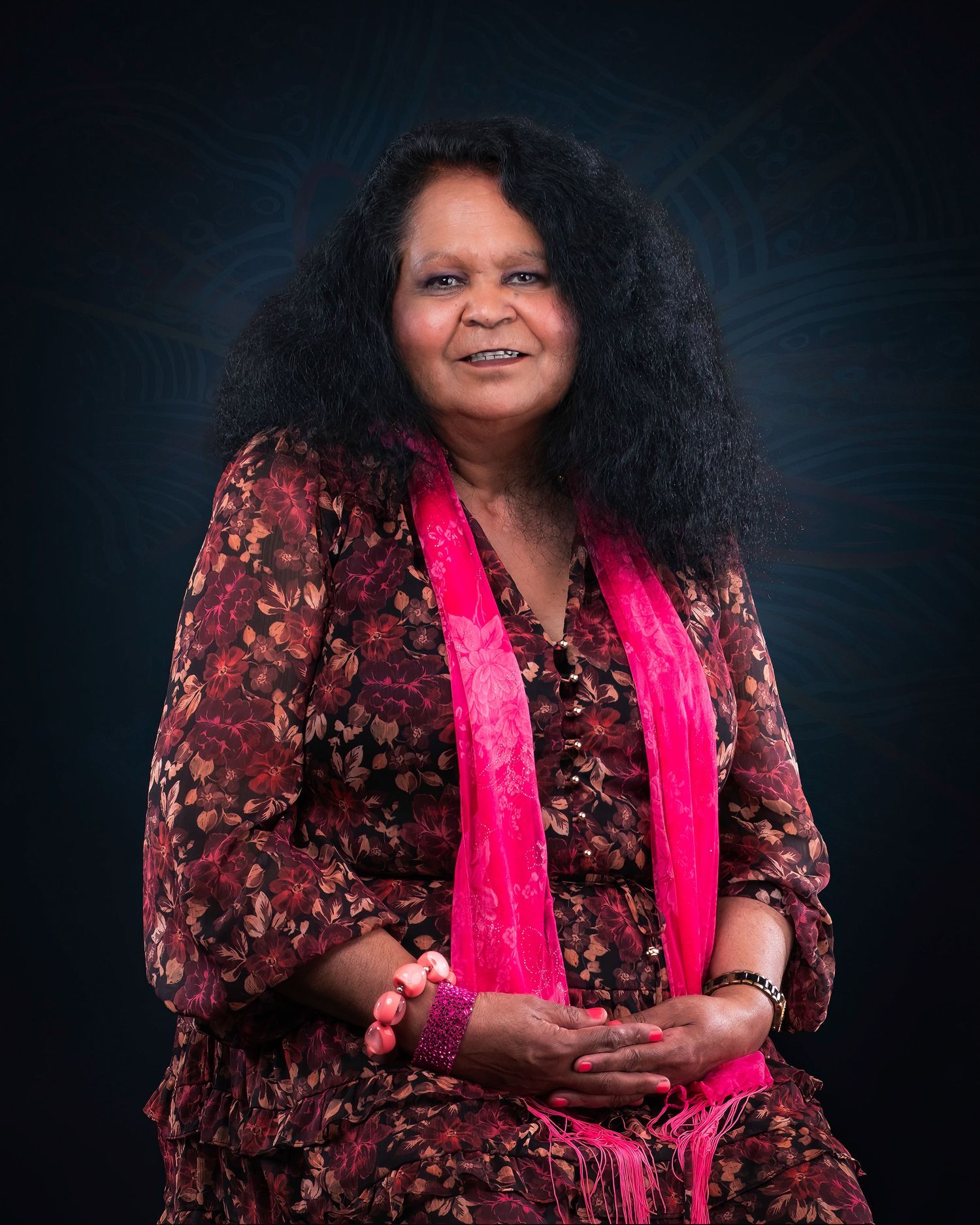 A person wearing a dark floral jacket and bright pink top poses against a dark studio backdrop.