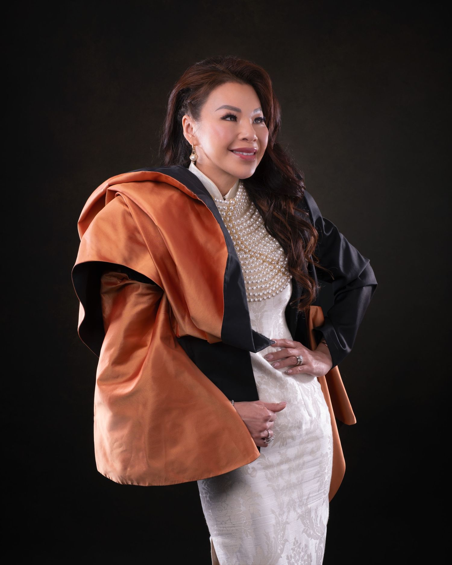 A person poses against a dark background wearing a brown coat over a cream sweater and white dress in an elegant portrait.