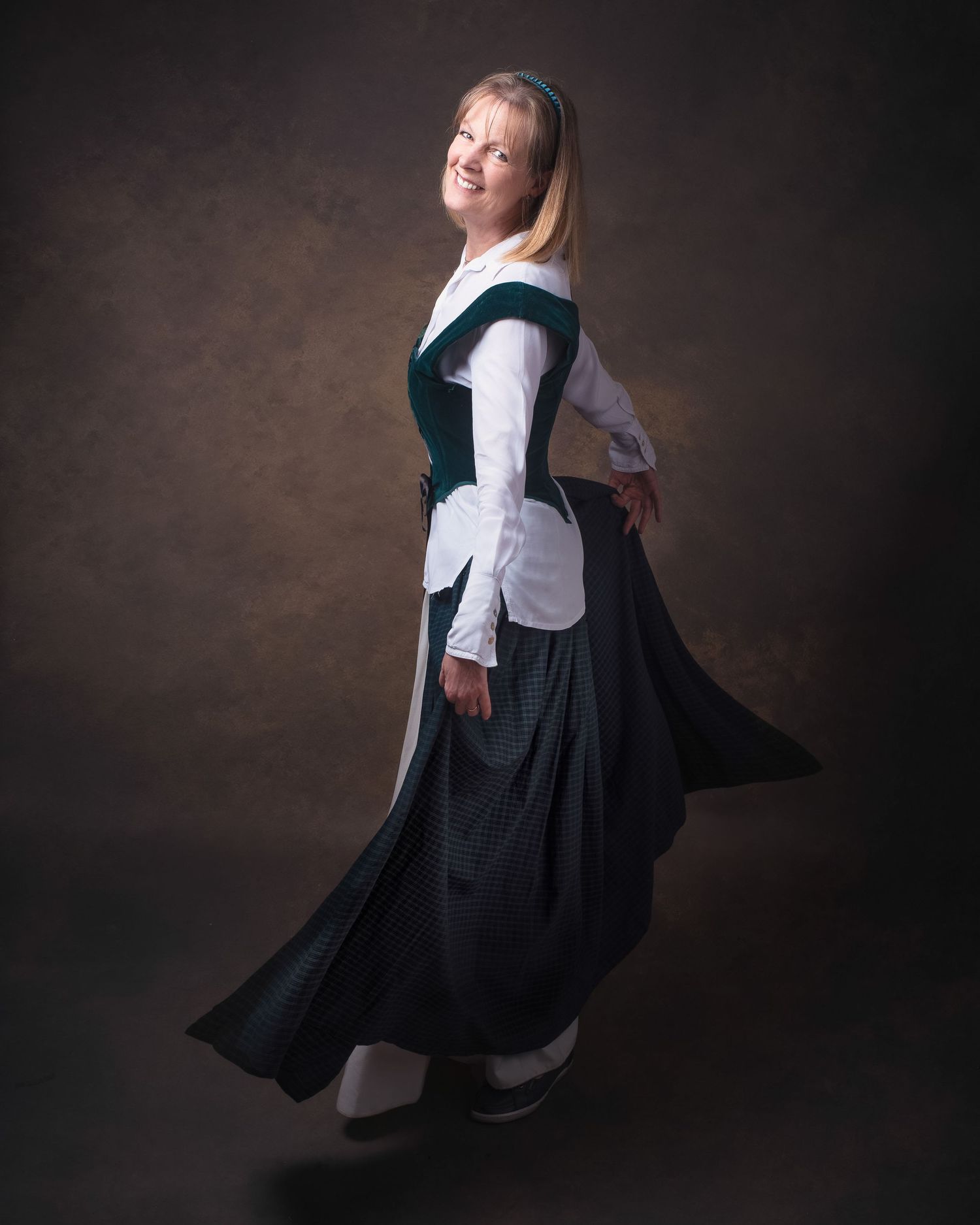 A person in a traditional maid's uniform with long skirt and apron poses gracefully against a dark studio backdrop.