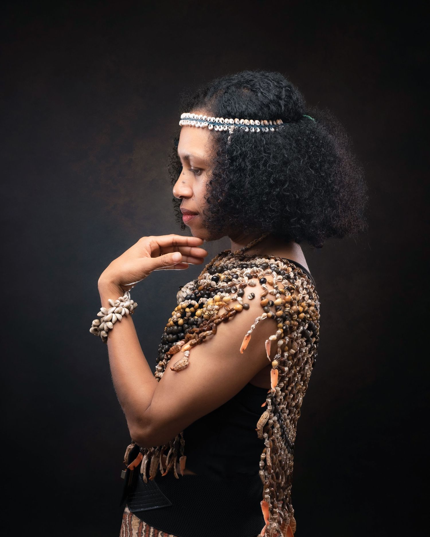 Studio portrait of person in glamorous jeweled accessories and formal attire with natural curly hair against dark background.