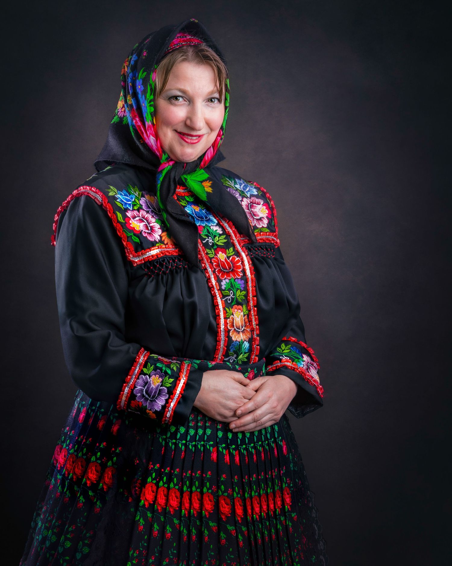 Traditional Eastern European folk costume with colorful floral headscarf, black dress with embroidered trim and pleated skirt.