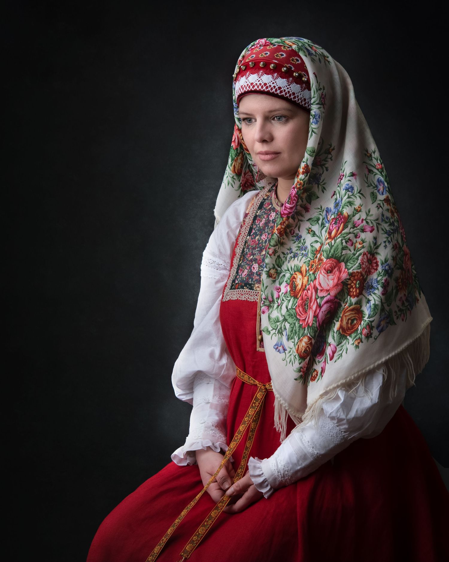 Traditional Russian folk costume with ornate floral headscarf, white blouse and red sarafan dress against dark background.