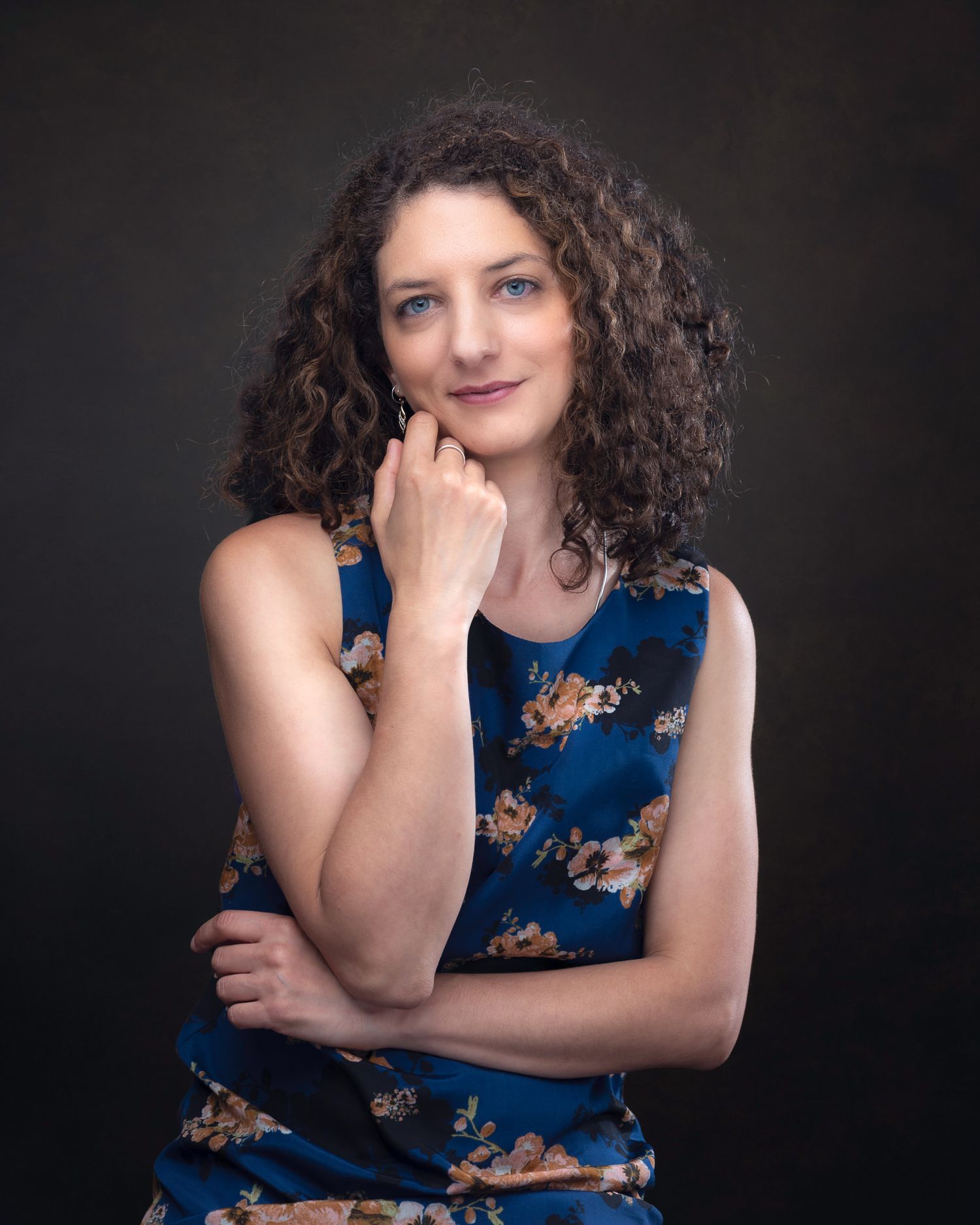 Studio portrait of a person in a navy floral dress with curly hair against a dark background, striking a thoughtful pose.