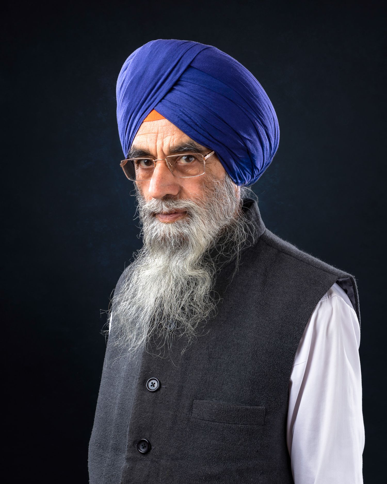 A person wearing a royal blue turban and glasses in a formal black vest and white shirt against a dark background.