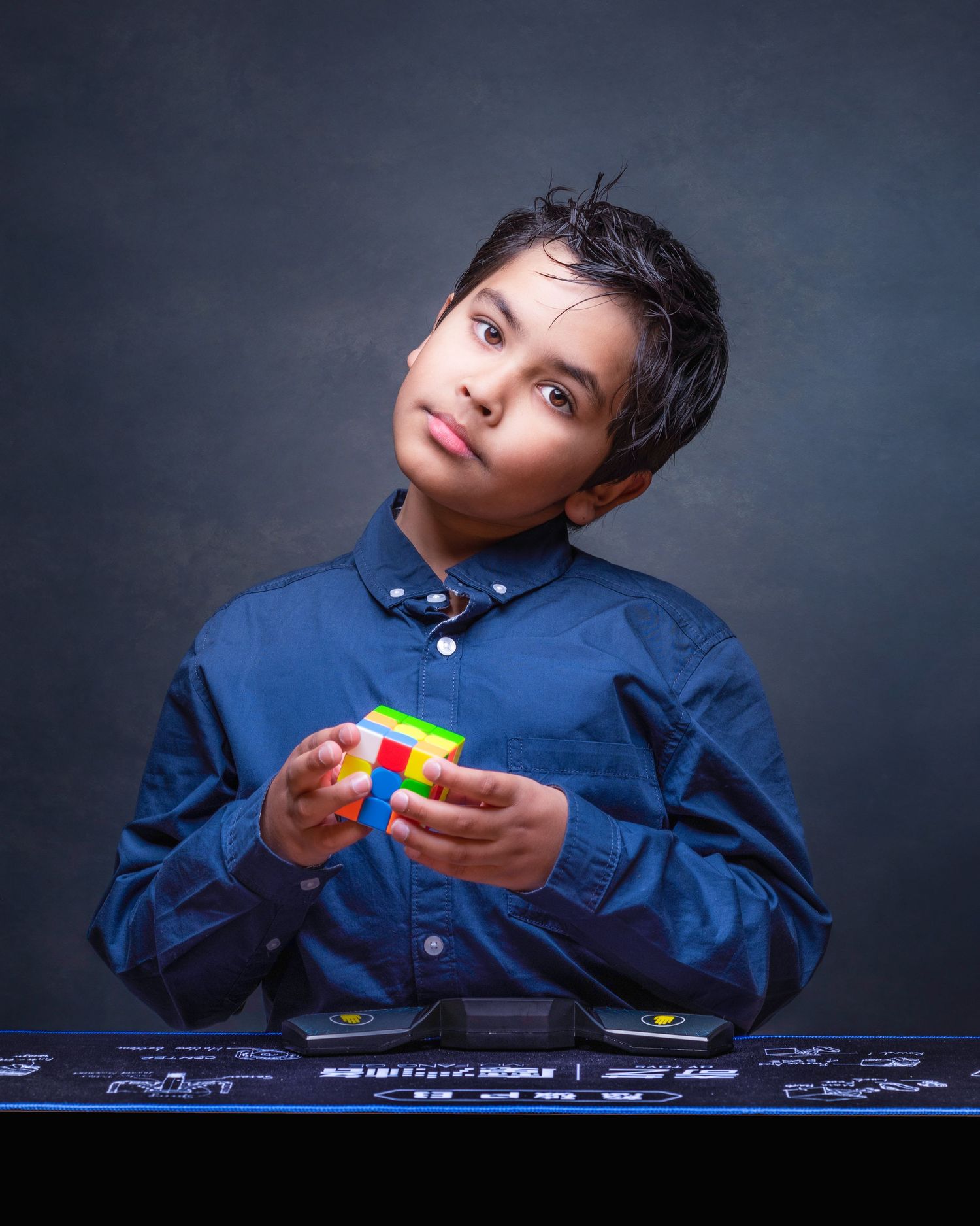 Young person in a blue button-down shirt holds a Rubik's cube puzzle while looking upward against a dark background.