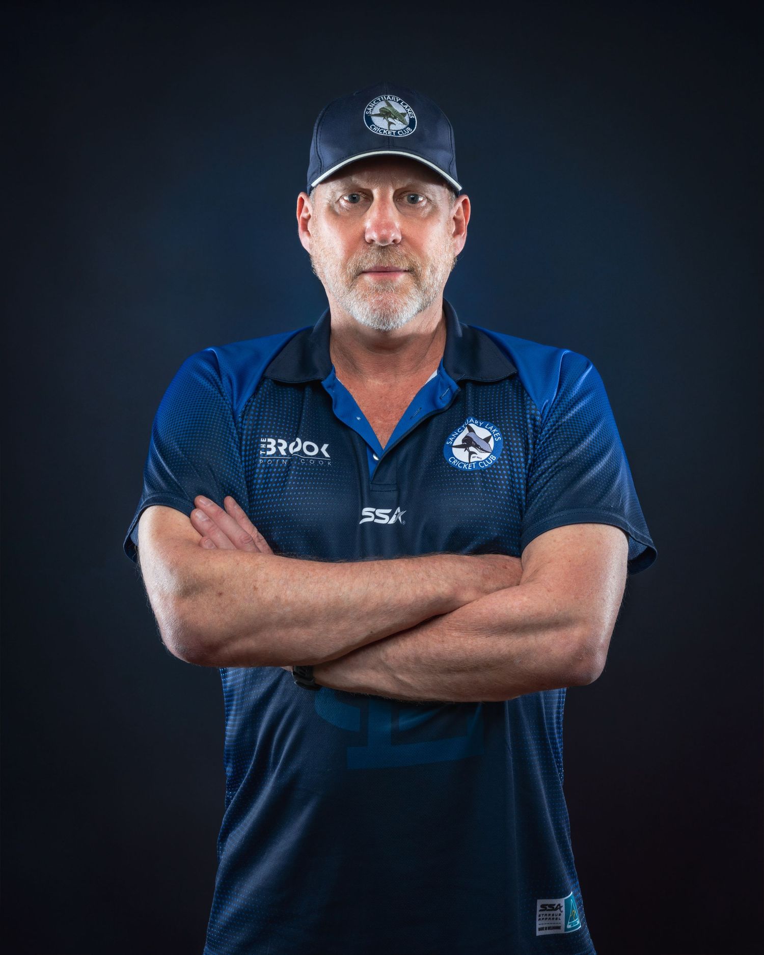 Coach wearing navy blue uniform with team logo and hat stands confidently with arms crossed against dark background.