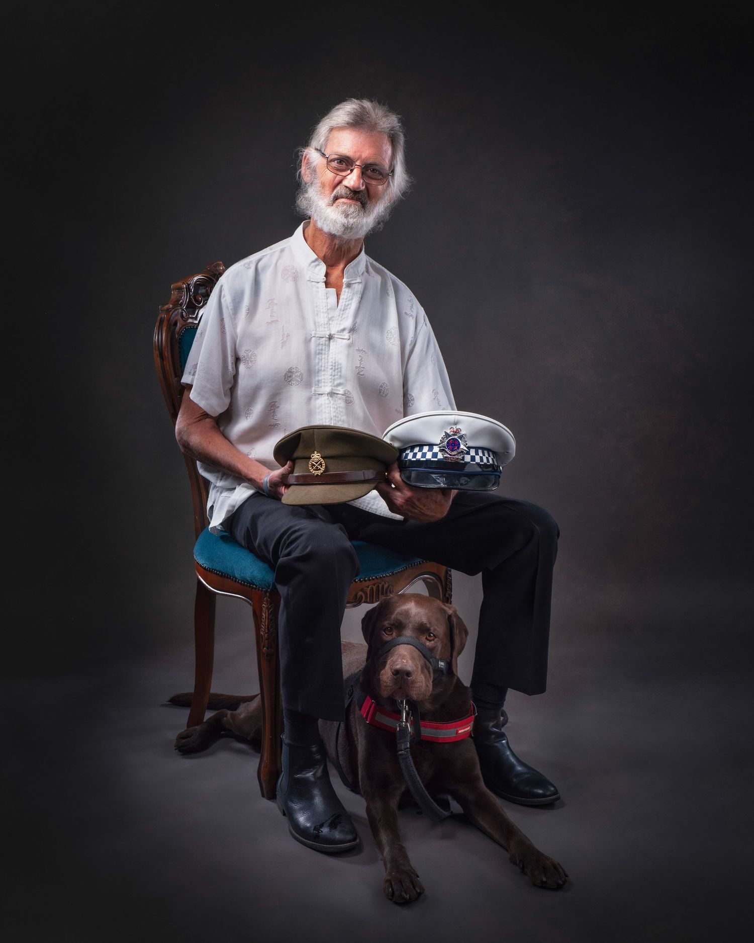 Portrait in a studio setting showing a person in white shirt seated on a chair with a dog at their feet and holding police hats.