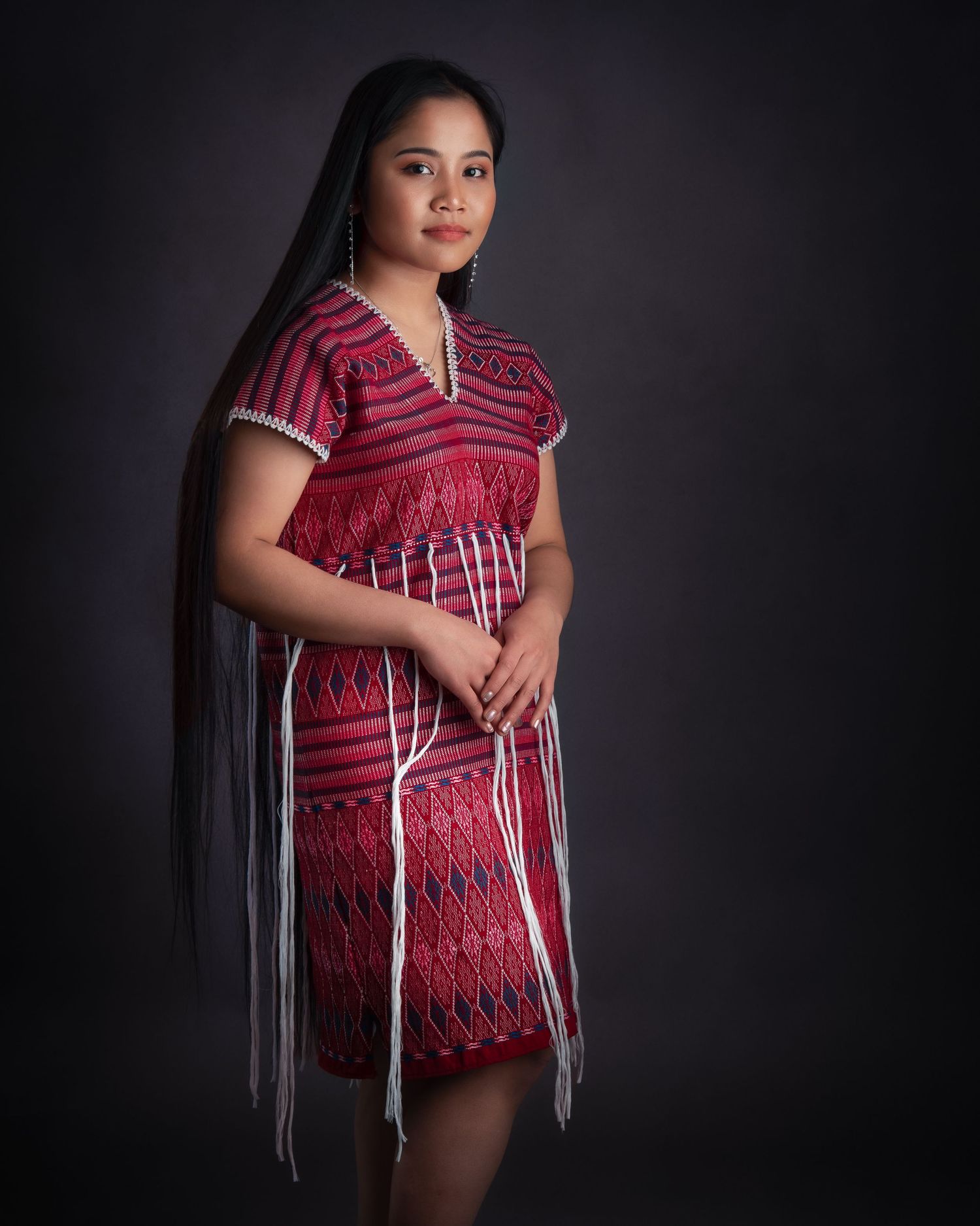 Modern traditional dress in red and white with geometric patterns and long fringe details against dark background in studio setting.