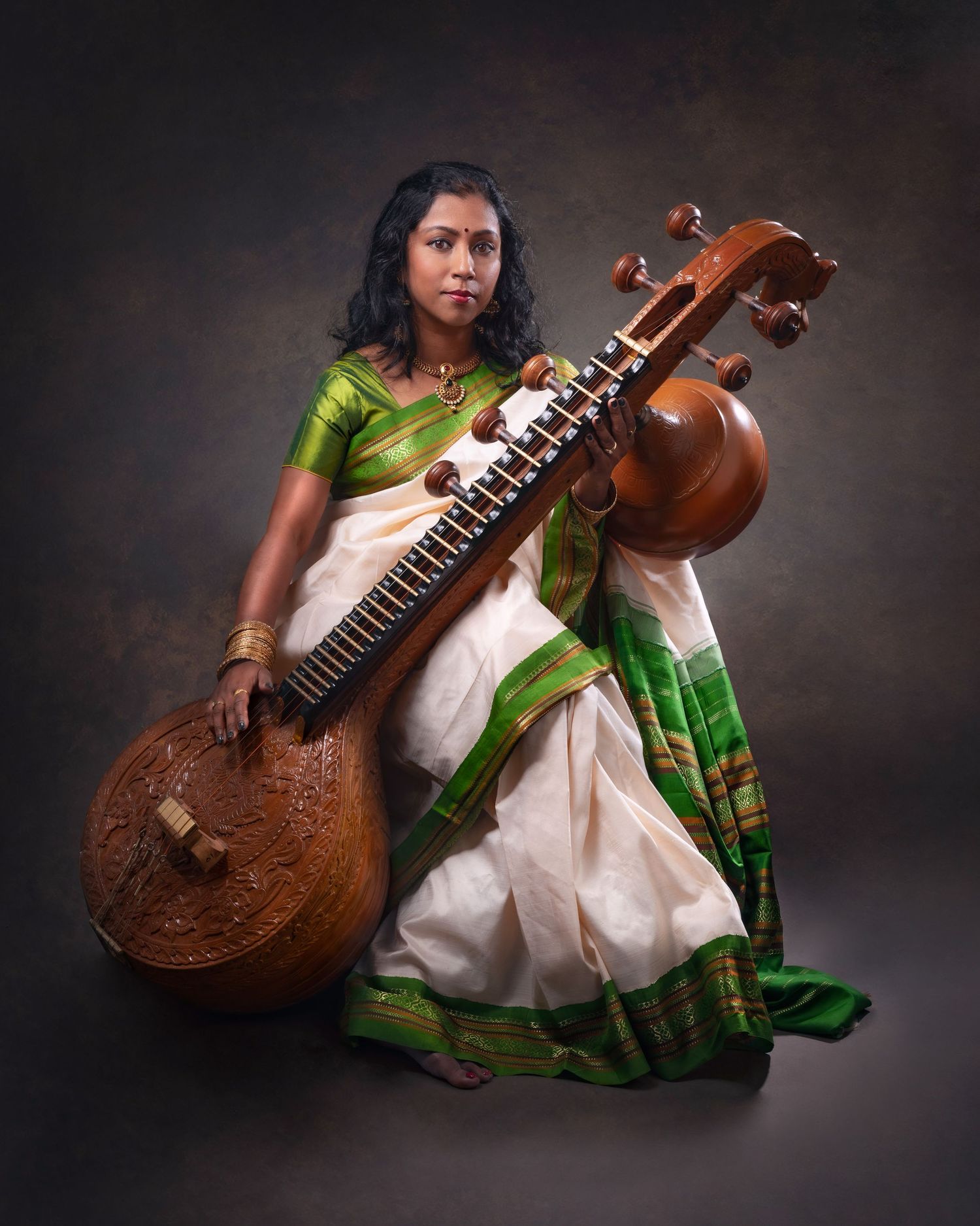 A musician in traditional attire plays the Saraswati Veena, an Indian classical string instrument, against a dark backdrop.