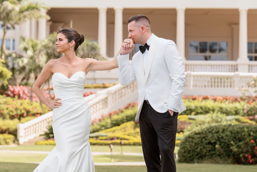 A couple in wedding attire dances together on a sunny patio surrounded by lush gardens and stone columns.