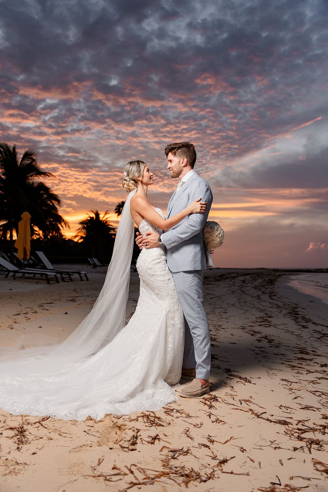 A couple shares a romantic moment on a beach at sunset with dramatic orange clouds.