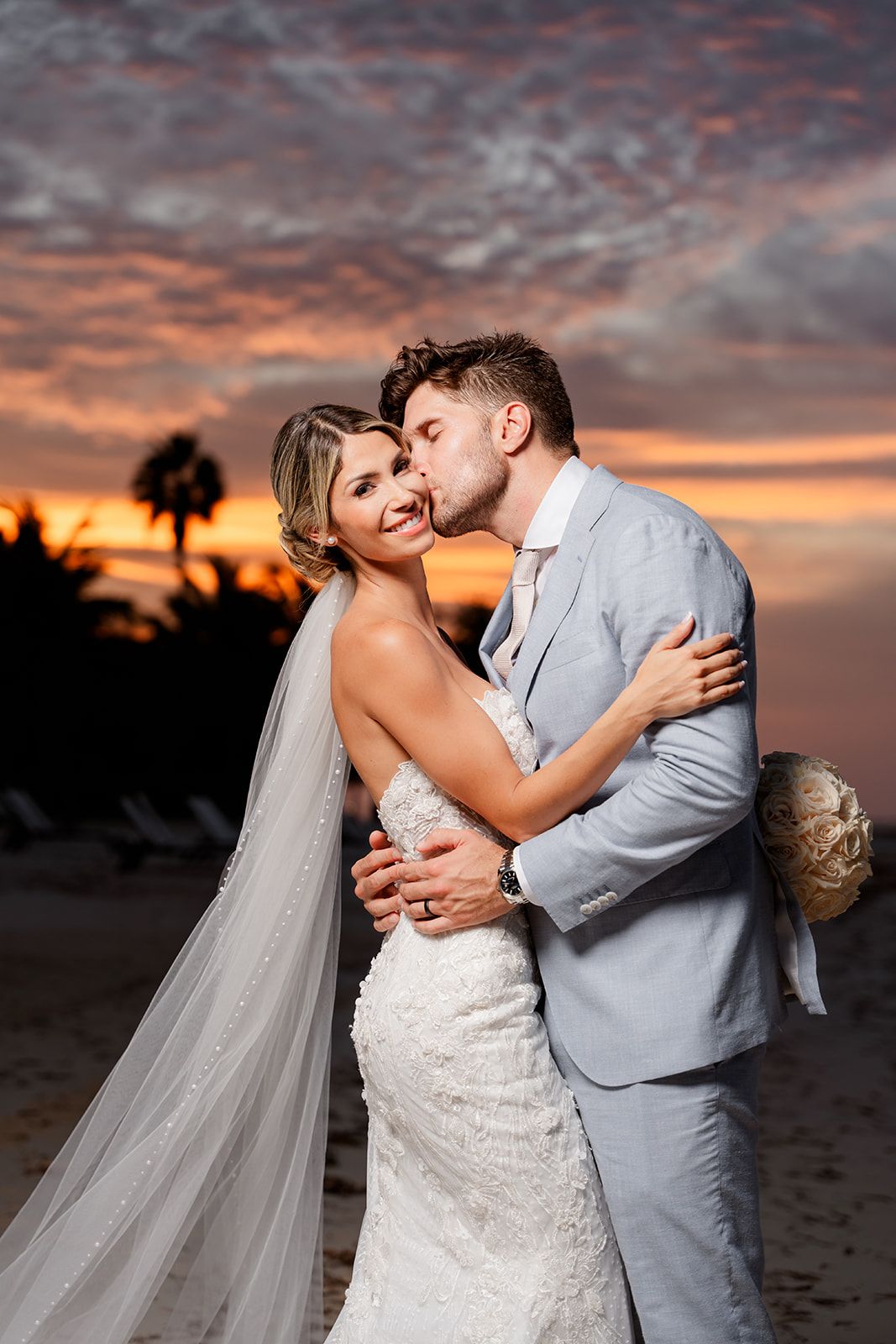 A romantic embrace between newlyweds on a beach during a vibrant sunset.
