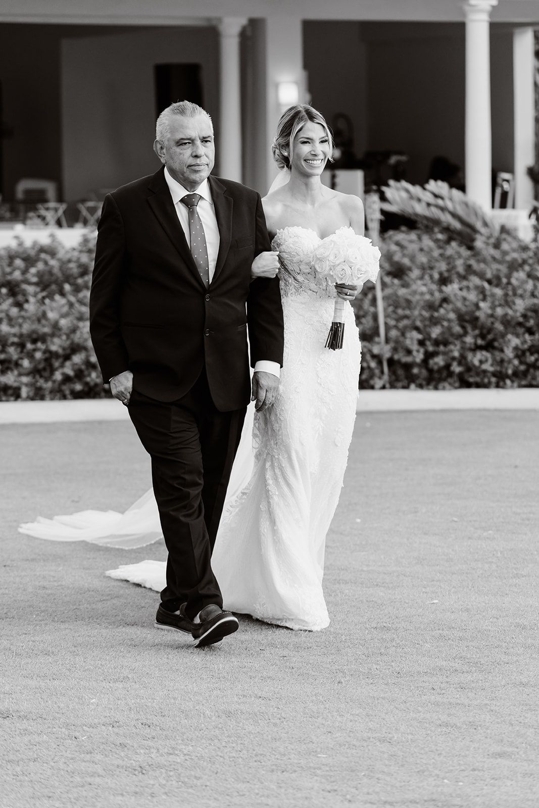 A bride in a white dress walks down an outdoor path with her escort on her wedding day.