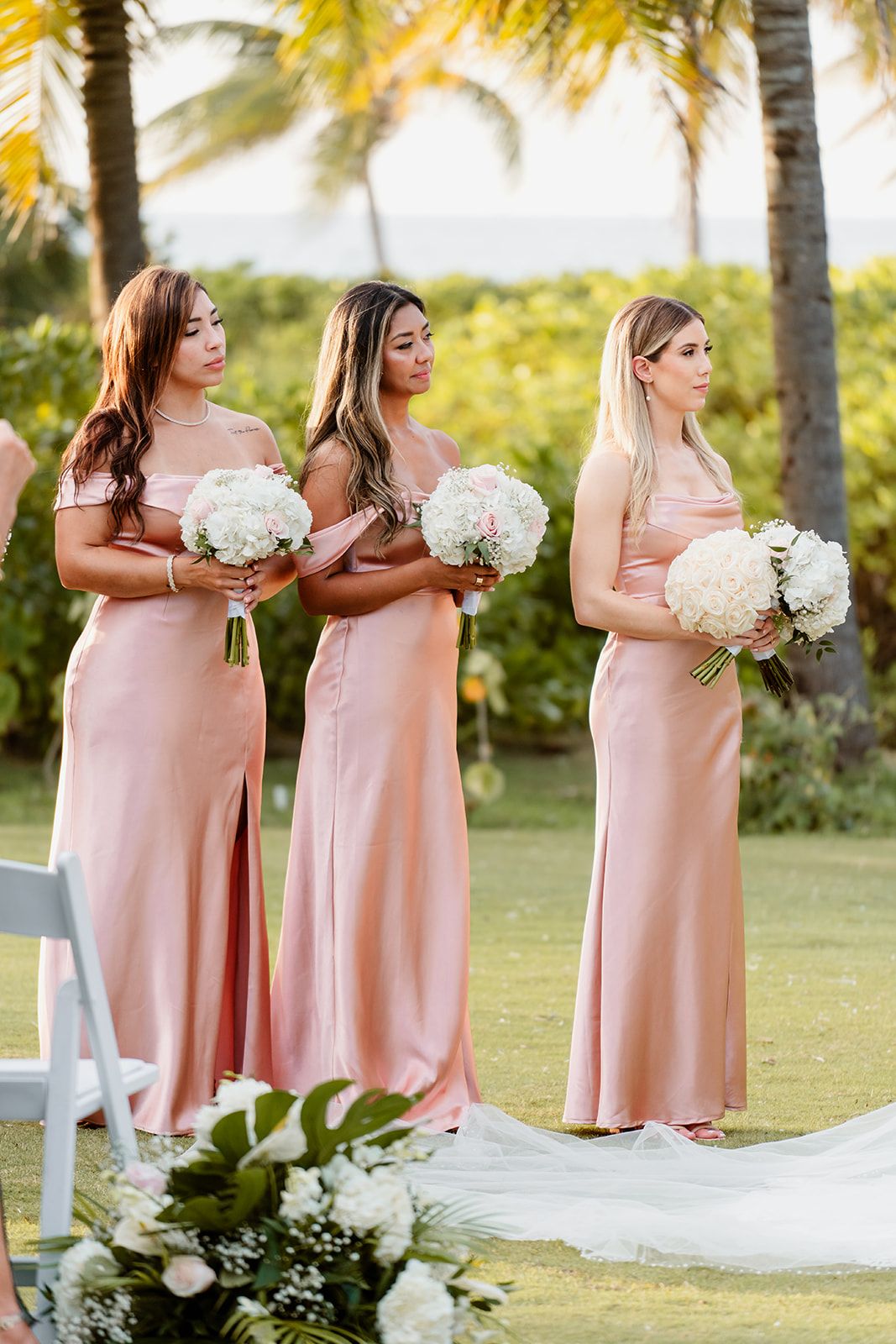 Three bridesmaids in pink dresses hold white bouquets during an outdoor tropical wedding ceremony.