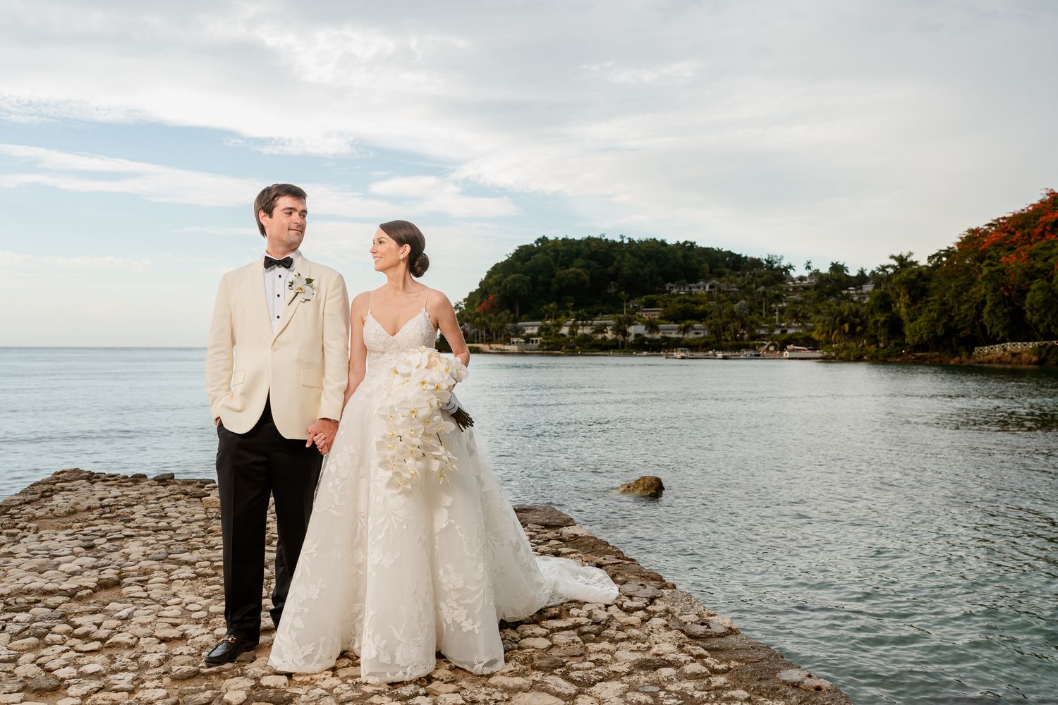 An elegant couple poses on a rocky beach at sunset with a tranquil bay and lush hillside in the background.