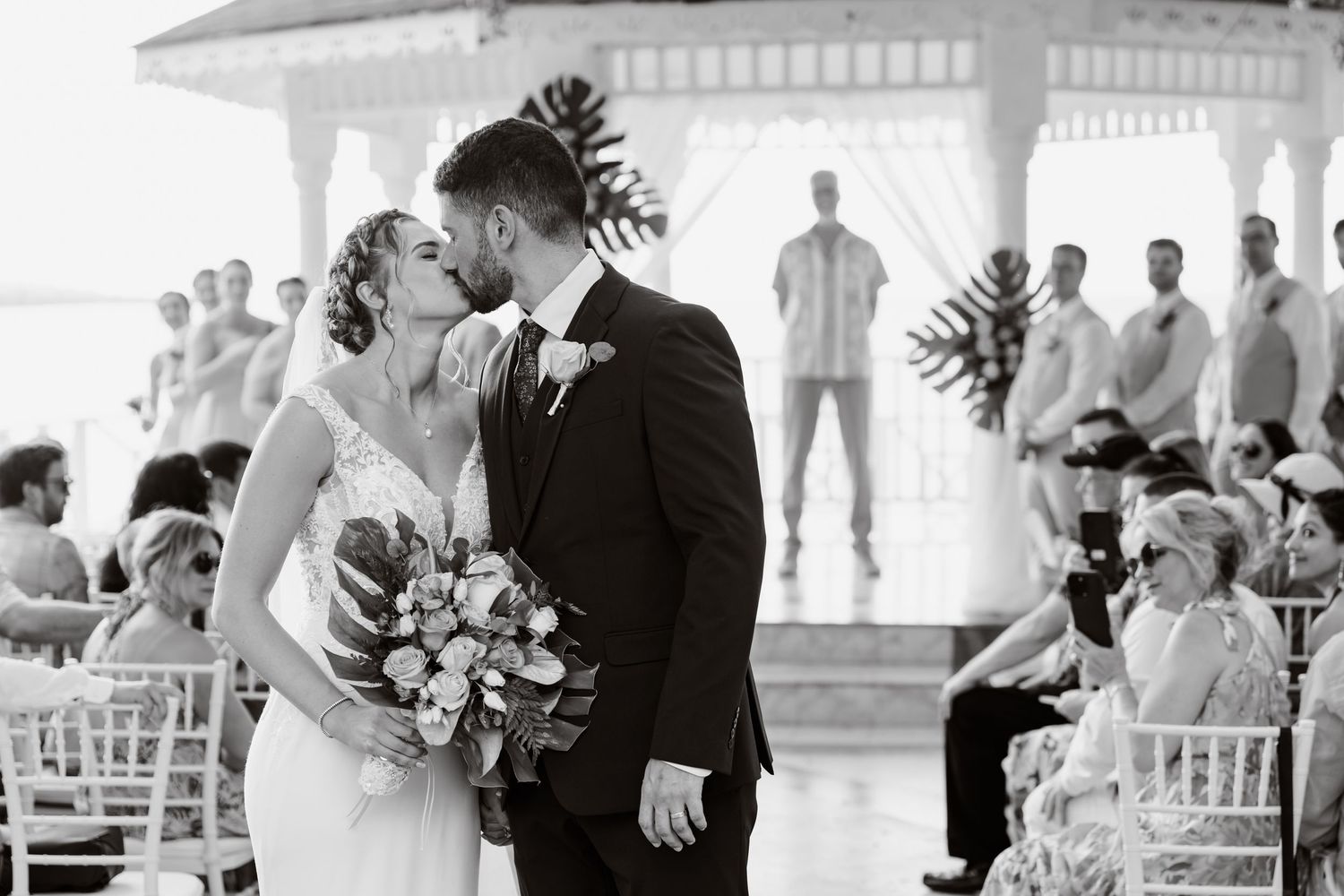 Black and white photograph showing a romantic moment during an outdoor wedding ceremony at a gazebo with guests watching.