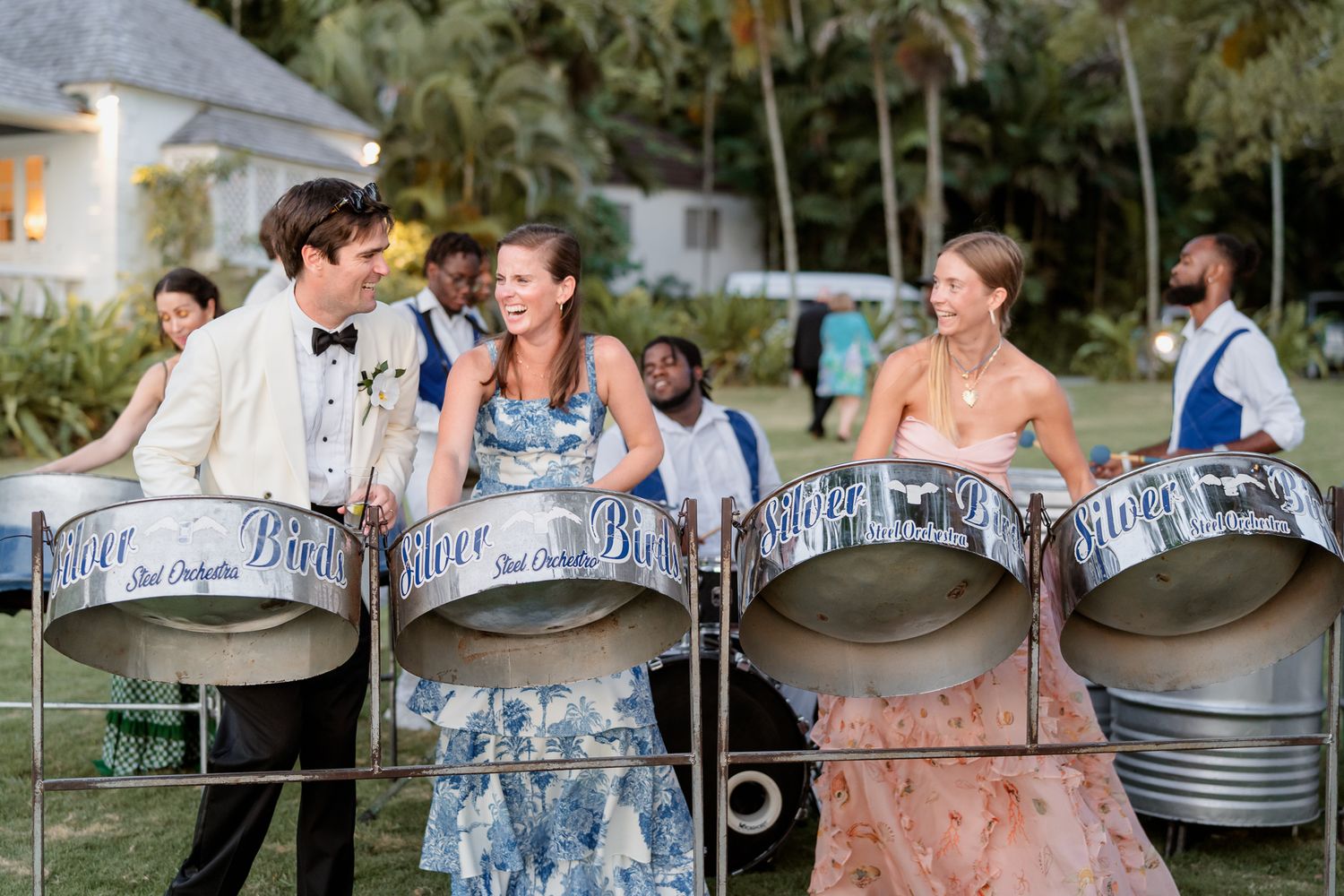 Steel drum musicians in formal attire perform at an outdoor event with palm trees in the background.