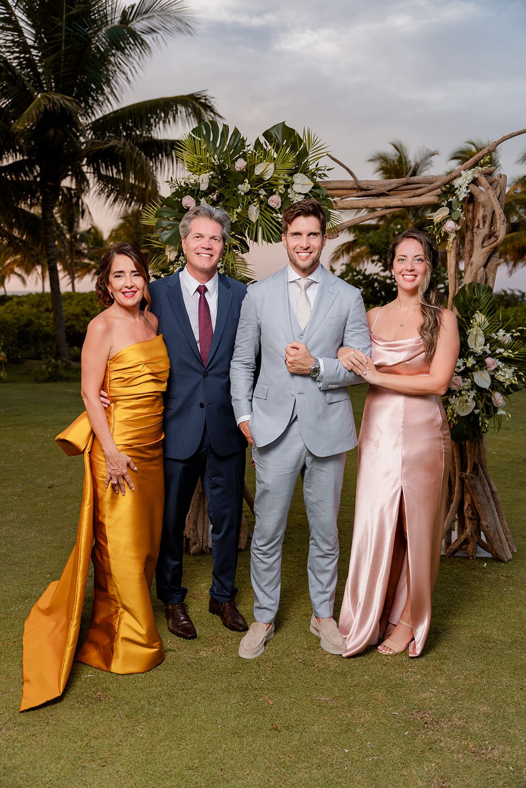 Wedding guests in formal attire pose together beneath a decorated arch with tropical palm trees.