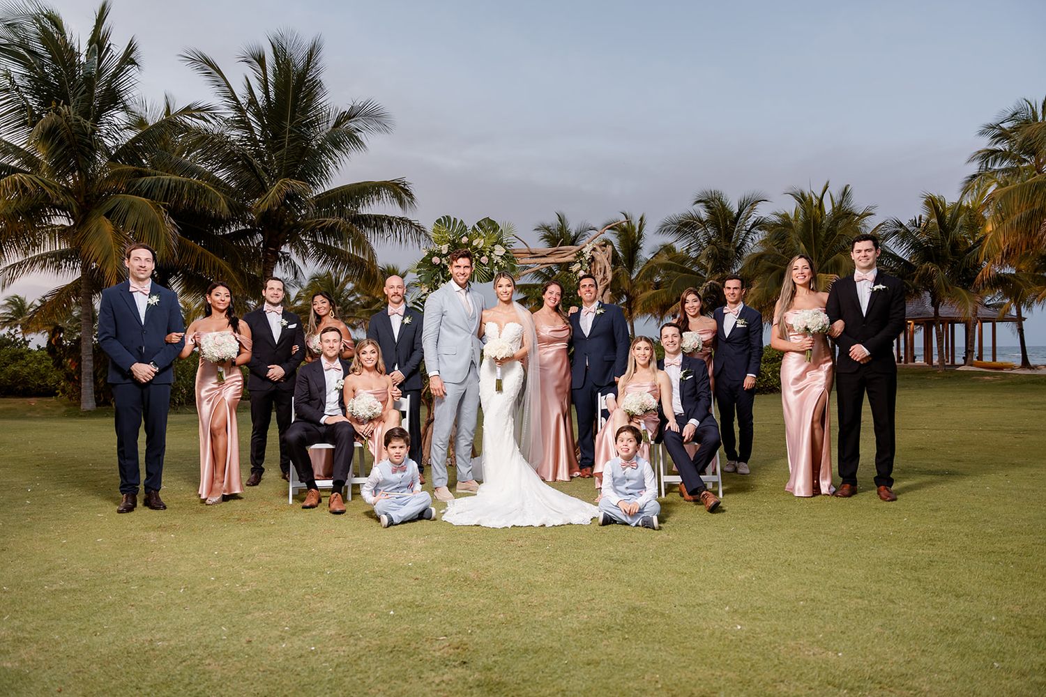 Wedding party and family members gather for a formal group photo on a tropical lawn with palm trees.