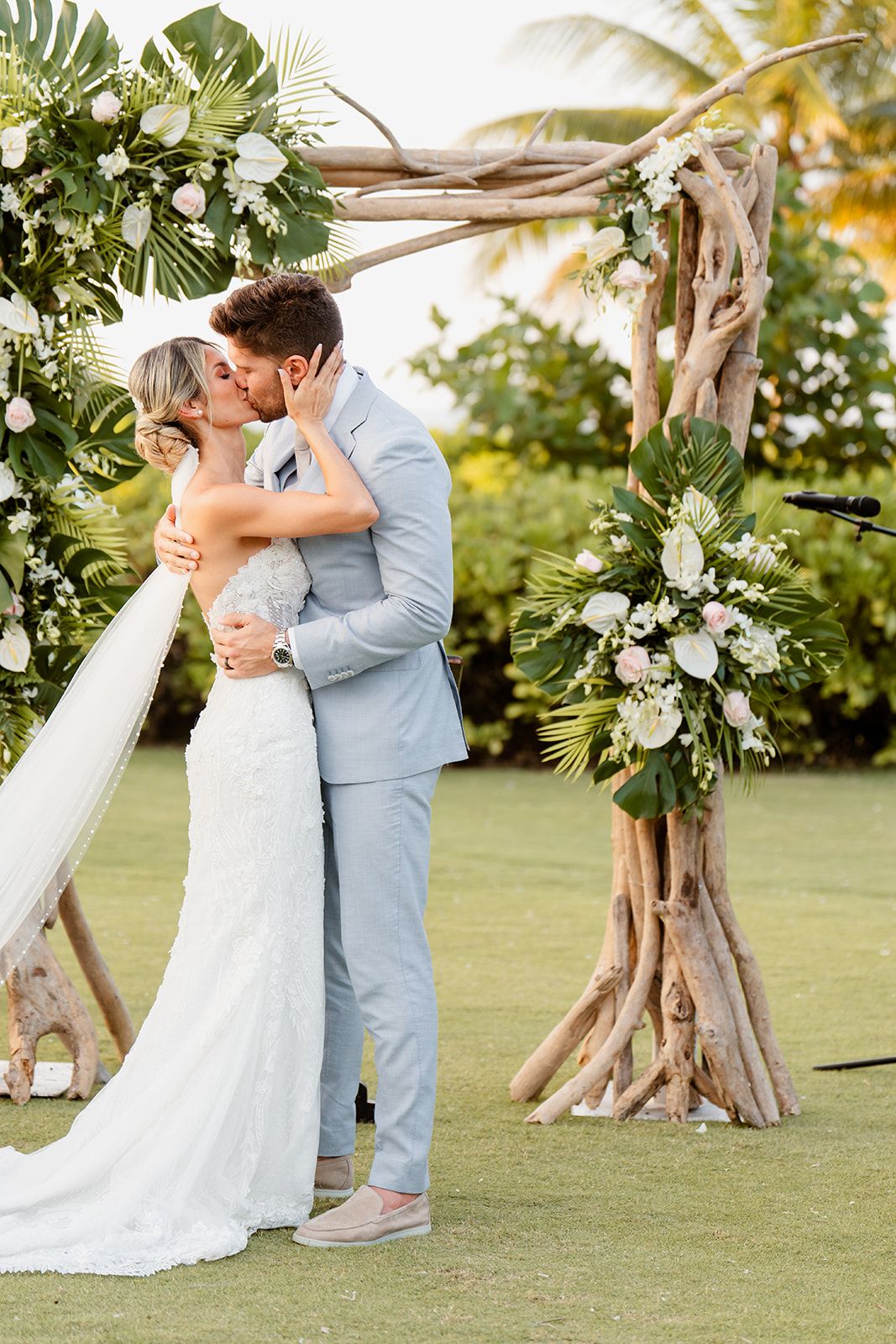 Newlyweds share their first kiss under a wooden arch decorated with tropical flowers and greenery.