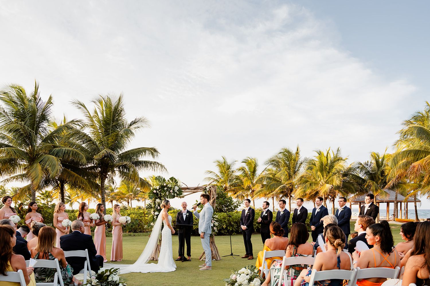 Wide shot of outdoor wedding ceremony under palm trees with seated guests viewing the couple.