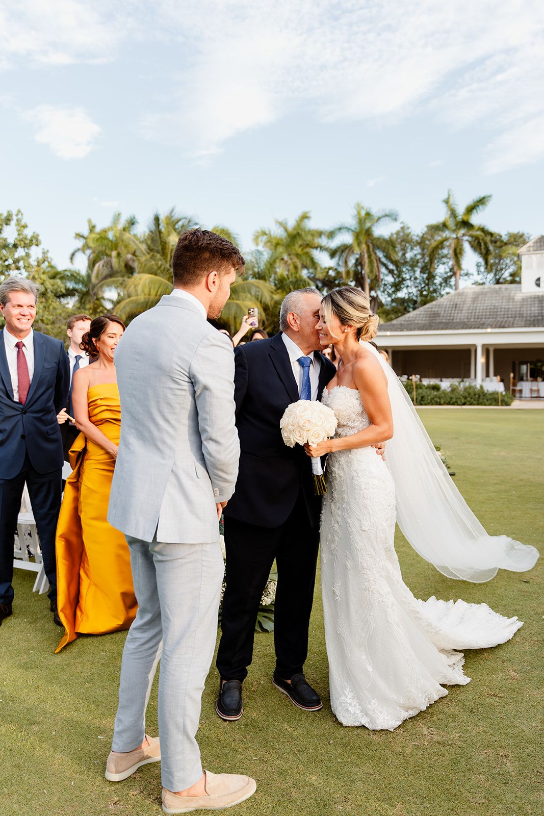 Wedding guests gather on a lush golf course surrounded by palm trees during a tropical ceremony.