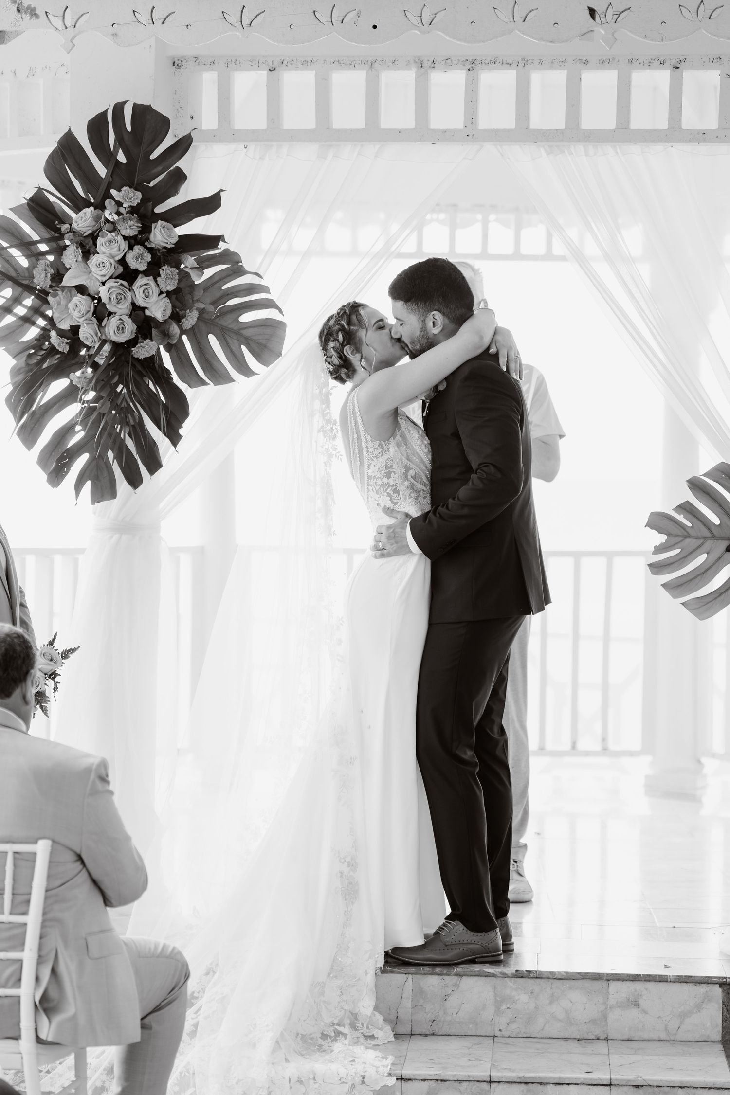 Black and white split image sequence showing couple embracing against white curtain backdrop with tropical monstera leaves.