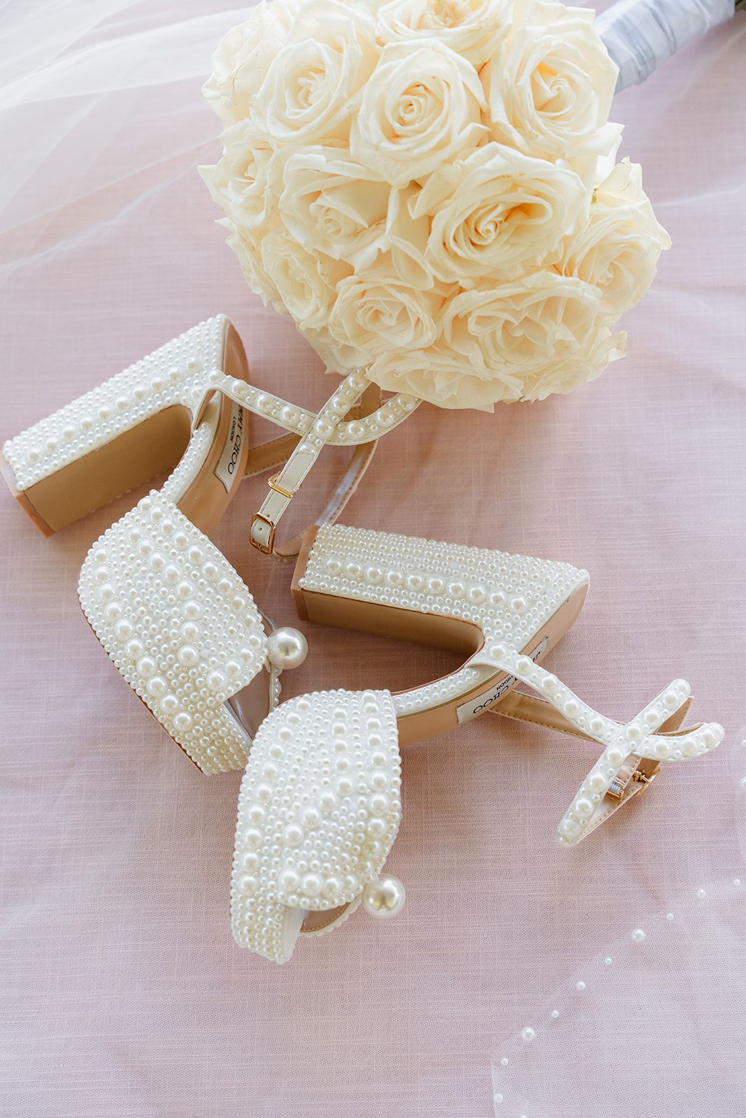 Elegant pearl-adorned bridal shoes next to a classic white rose bouquet on pink fabric.
