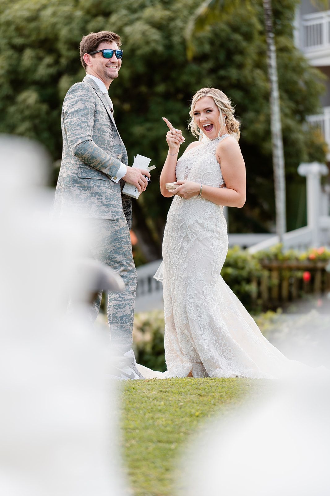 A couple shares a playful moment at an outdoor wedding, with the groom in military dress and bride in a lace gown.