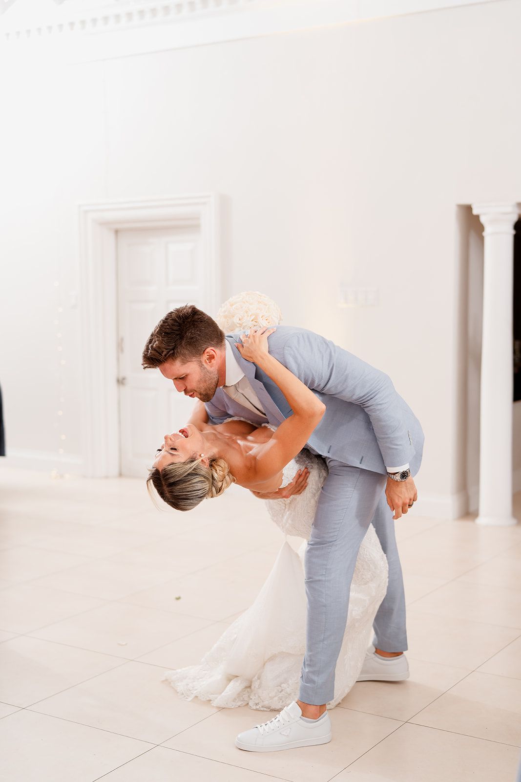 Bride and groom share playful dip dance pose in white reception venue.