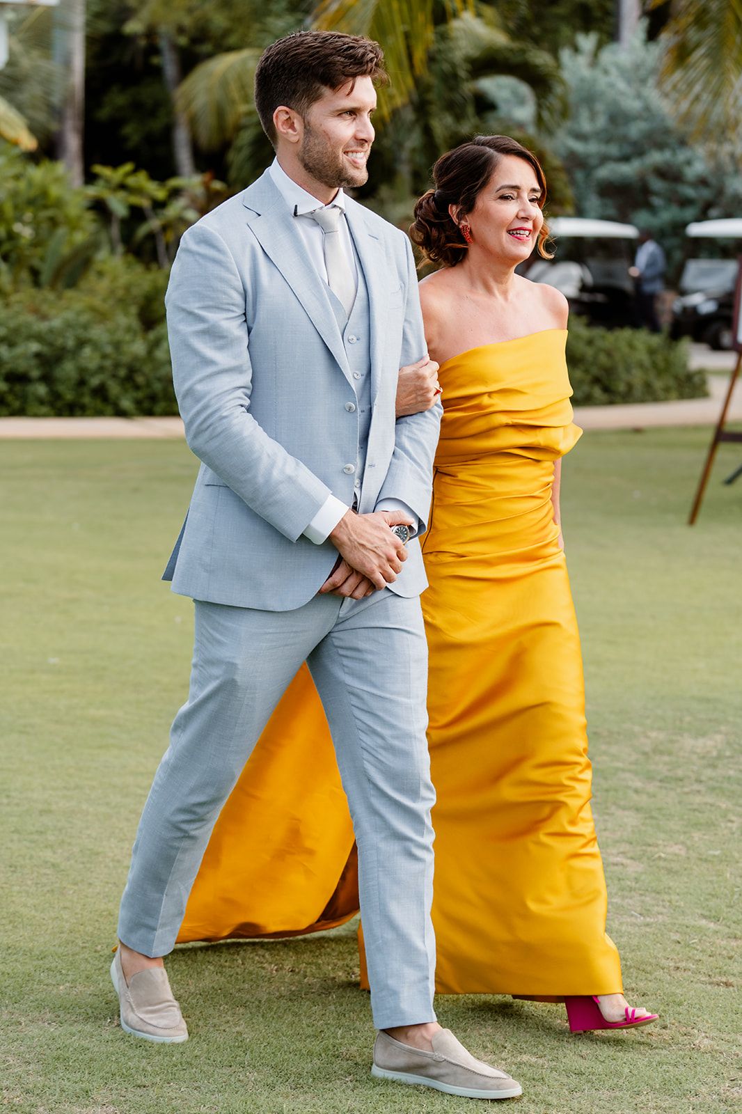 Groom in light blue suit walking with his mom in yellow dress on grass lawn.