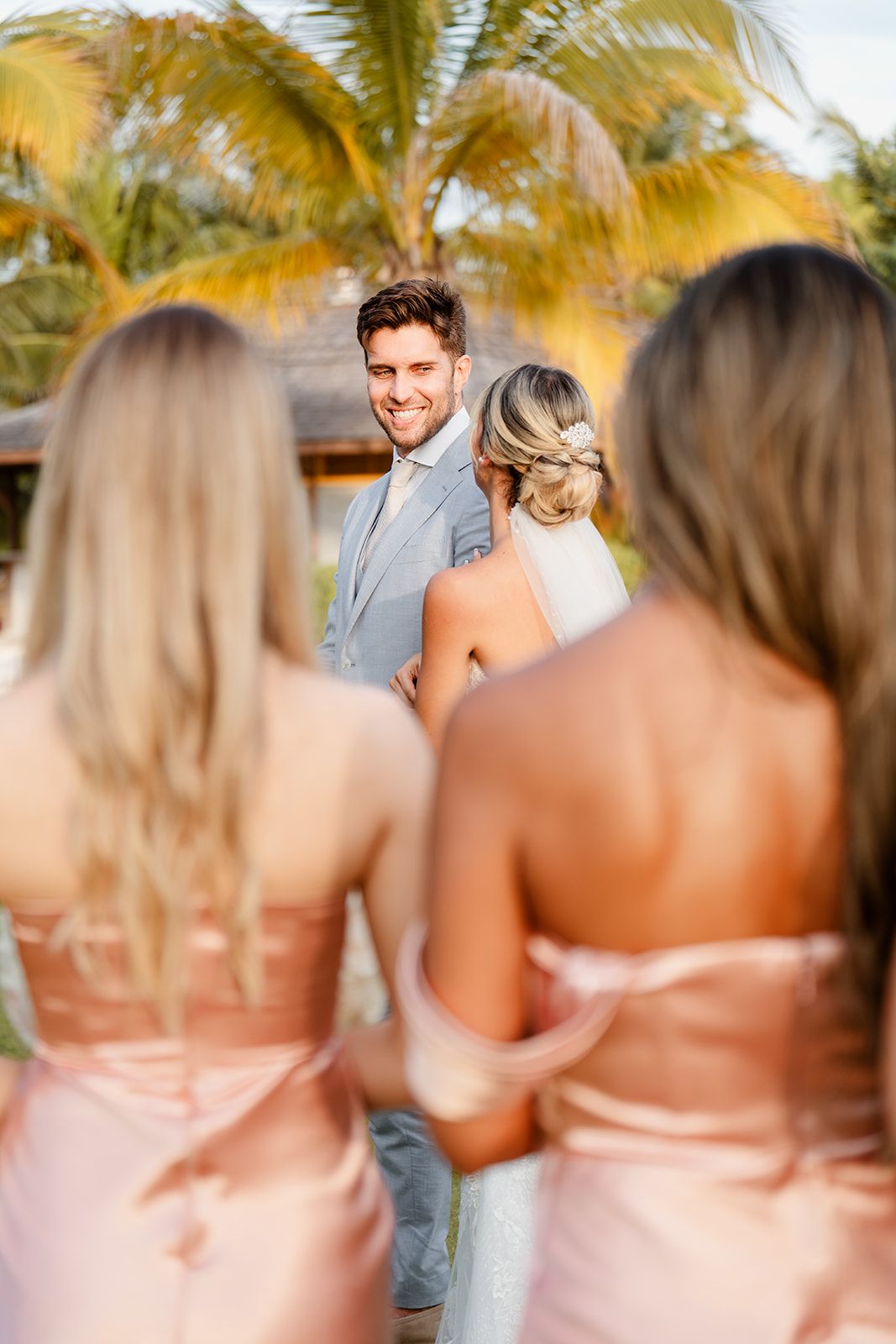 Wedding party members in pink dresses gather during a sunset tropical ceremony.
