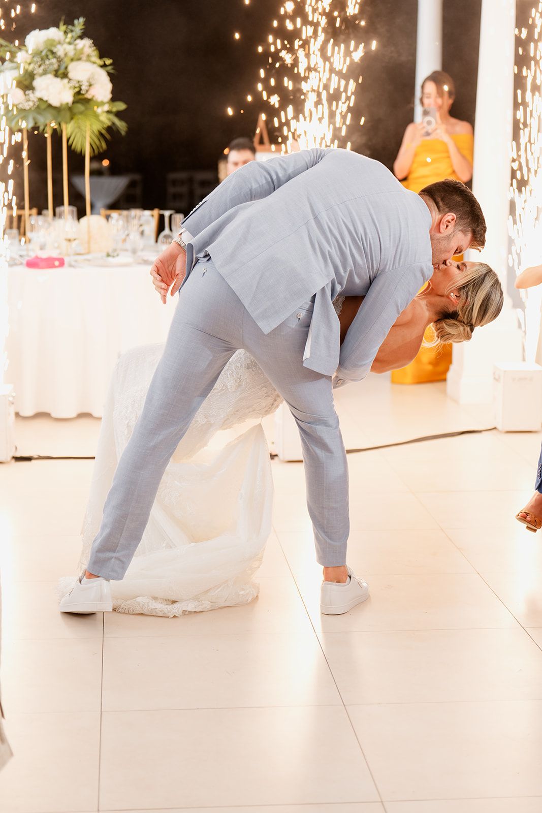 Groom in grey suit dips bride during wedding dance with sparklers in background.