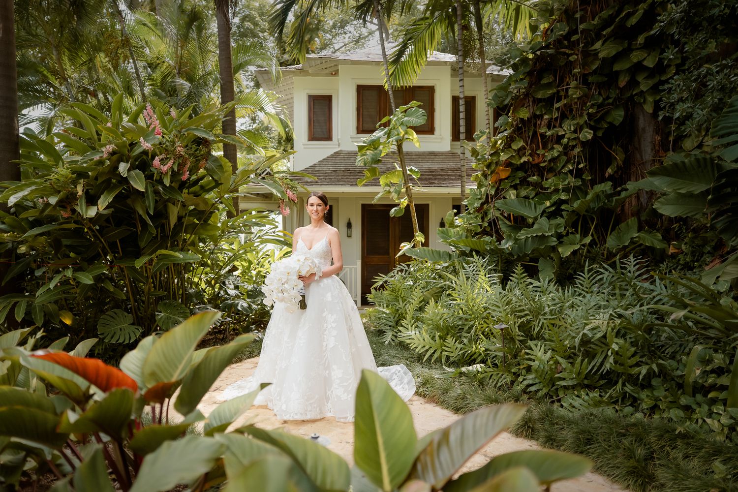 A white wedding dress amidst lush tropical garden foliage with a colonial-style house in the background.