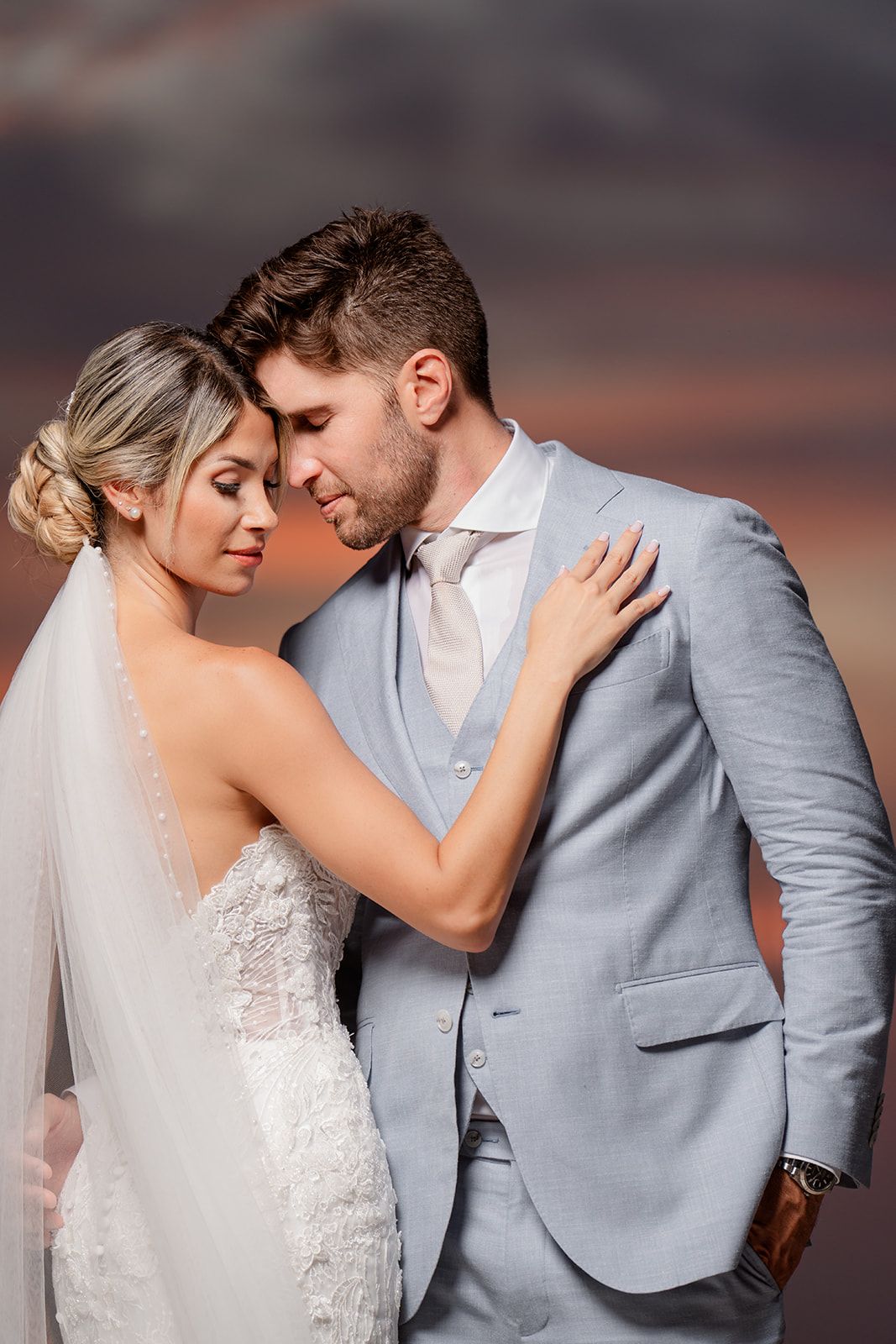 An intimate portrait of newlyweds in light gray suit and lace gown against sunset sky.