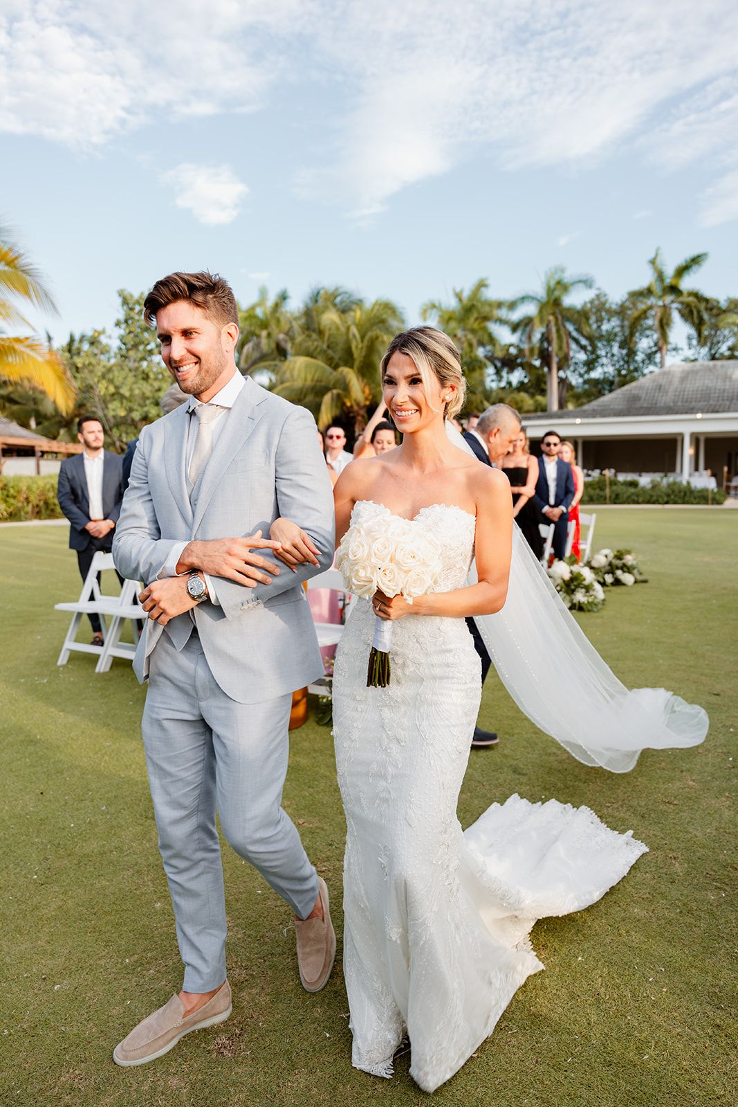 Happy newlyweds walking together on grass lawn after tropical outdoor wedding ceremony.