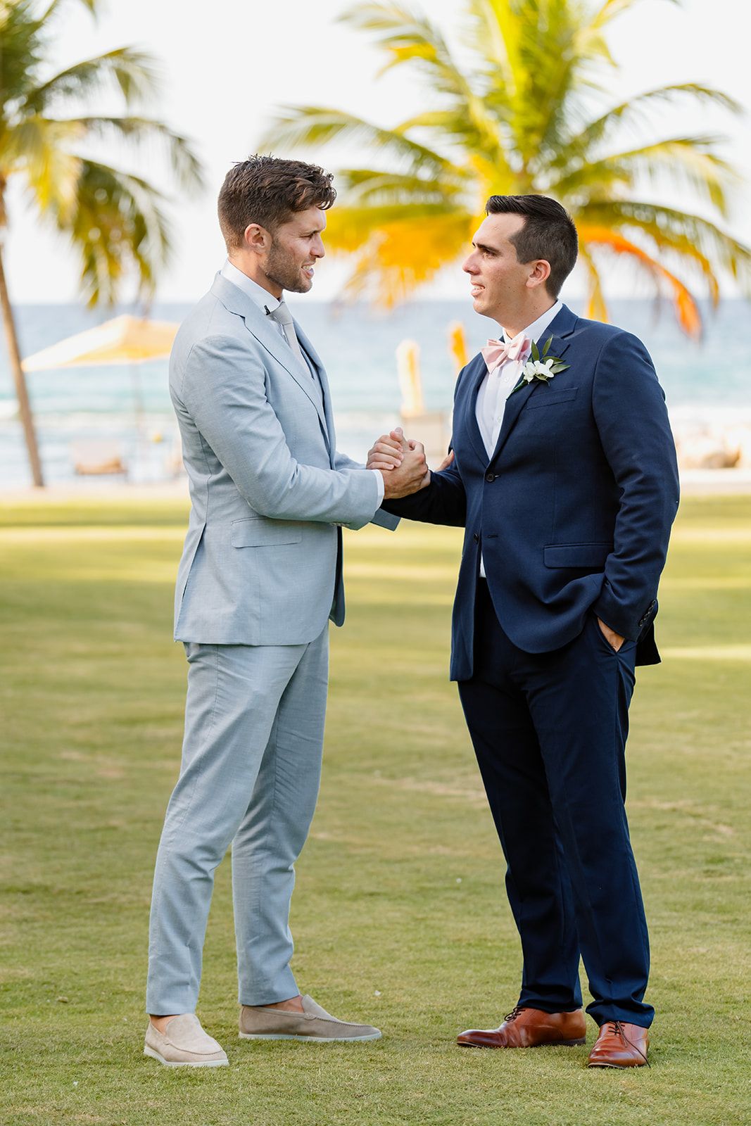 Two men in suits shake hands on a tropical beachfront lawn with palm trees.