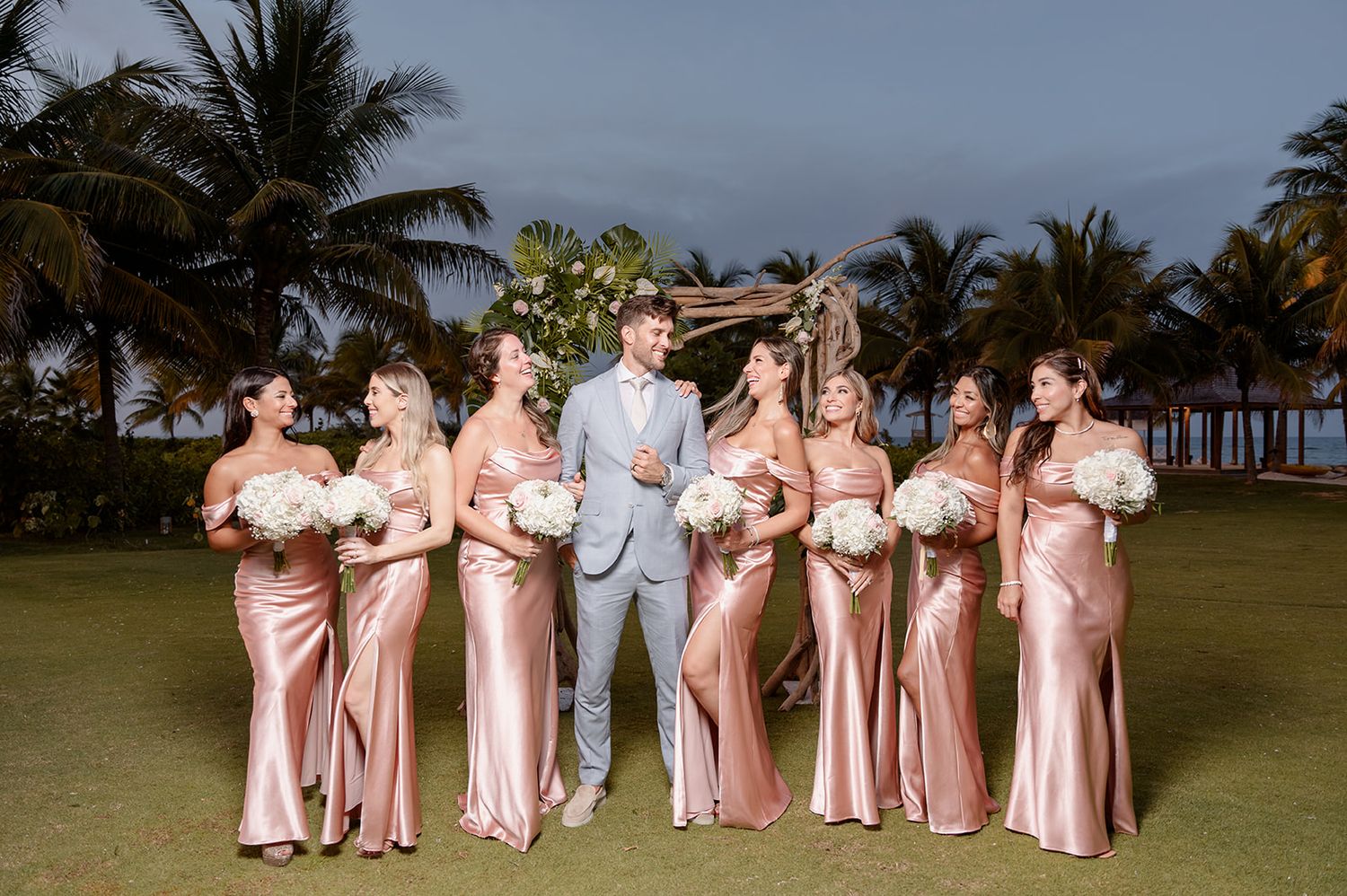 Bridesmaids in pink silk dresses pose with the groom on a palm-lined lawn.