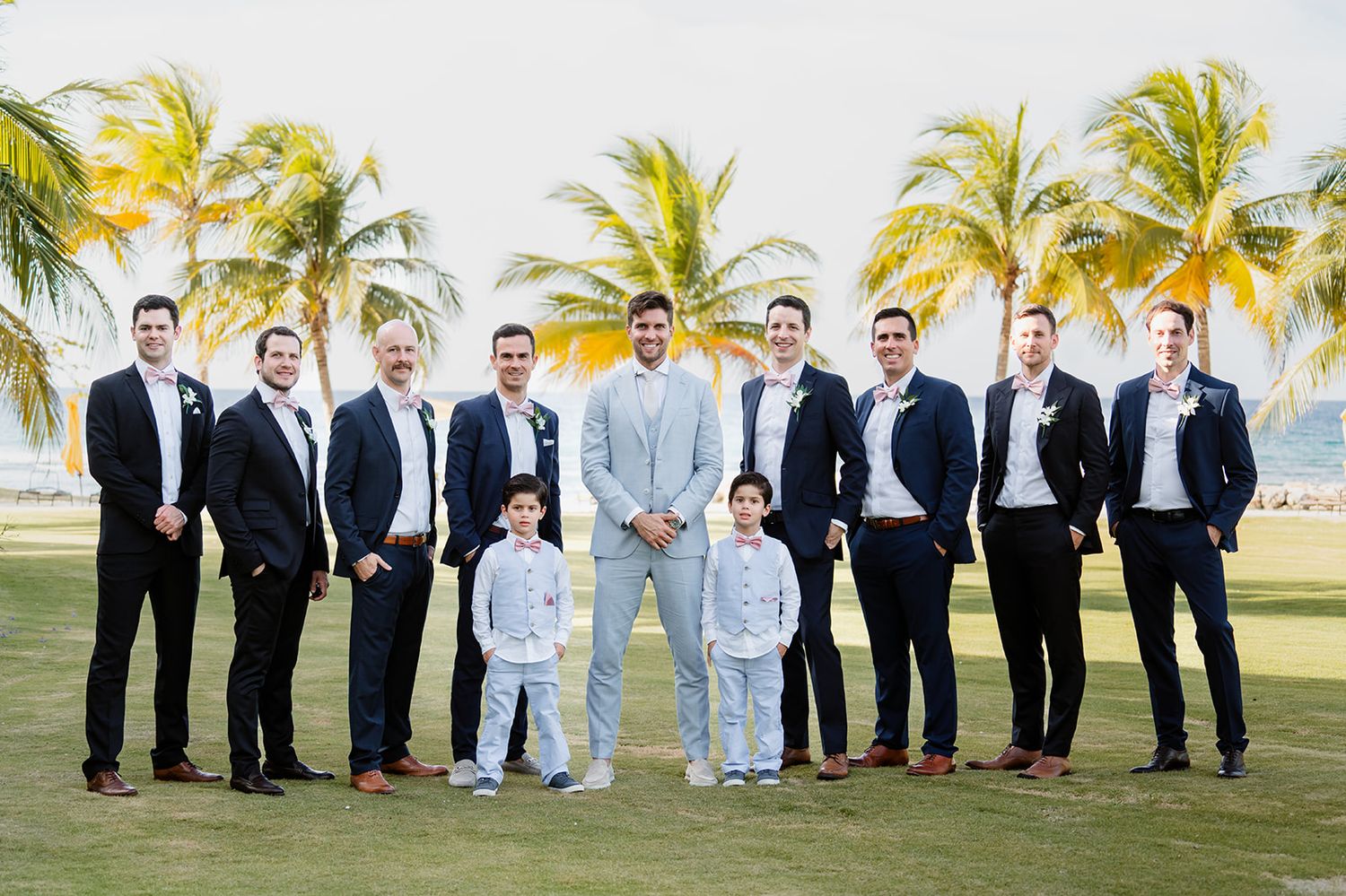 Groomsmen in dark suits stand with two young ring bearers in light suits on a tropical palm-lined lawn.
