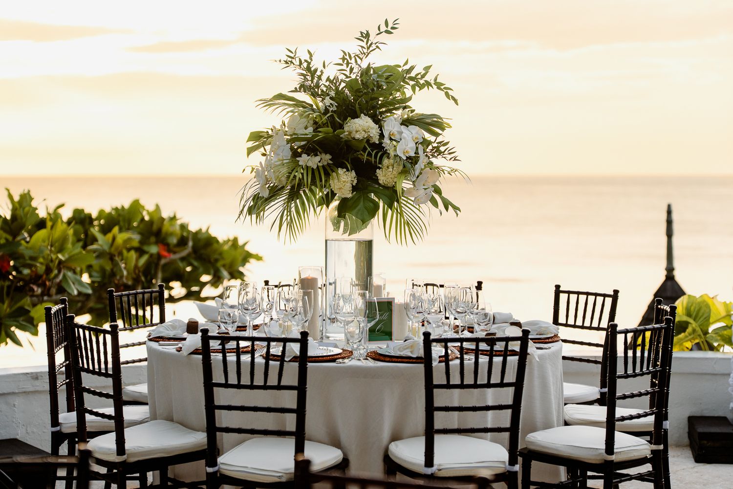 Elegant beachside wedding reception table with palm centerpiece and dark Chiavari chairs overlooking the ocean at sunset.