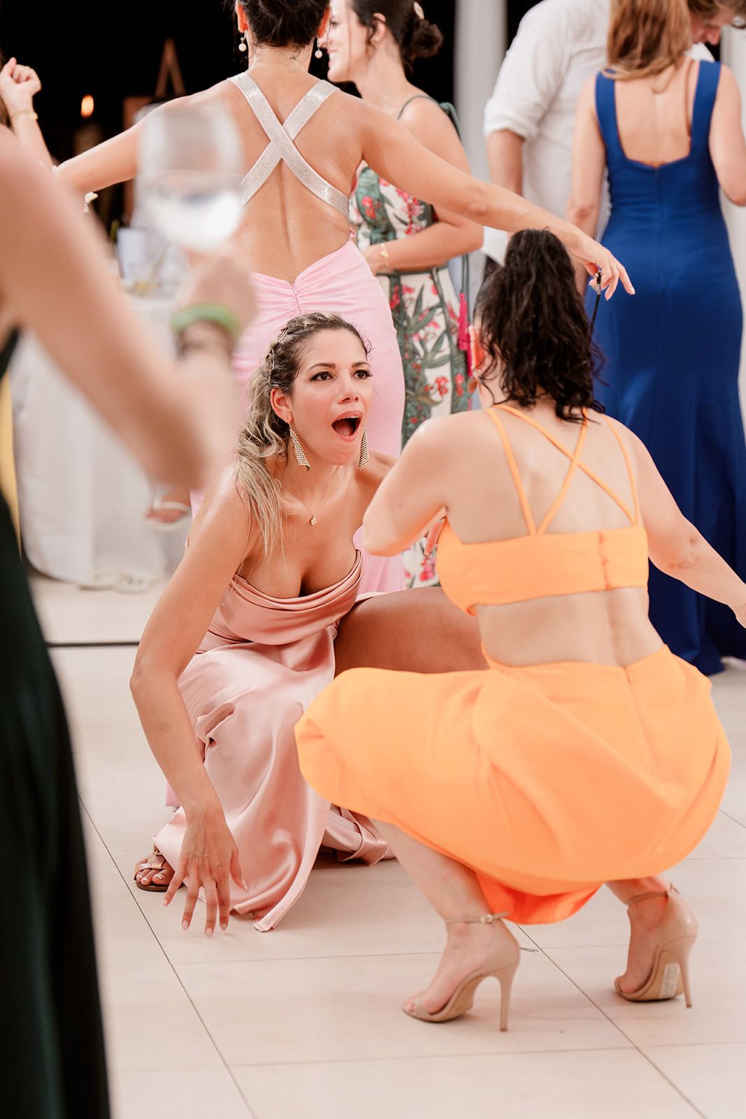 Wedding guests in colorful dresses dance and crouch low on a white dance floor.
