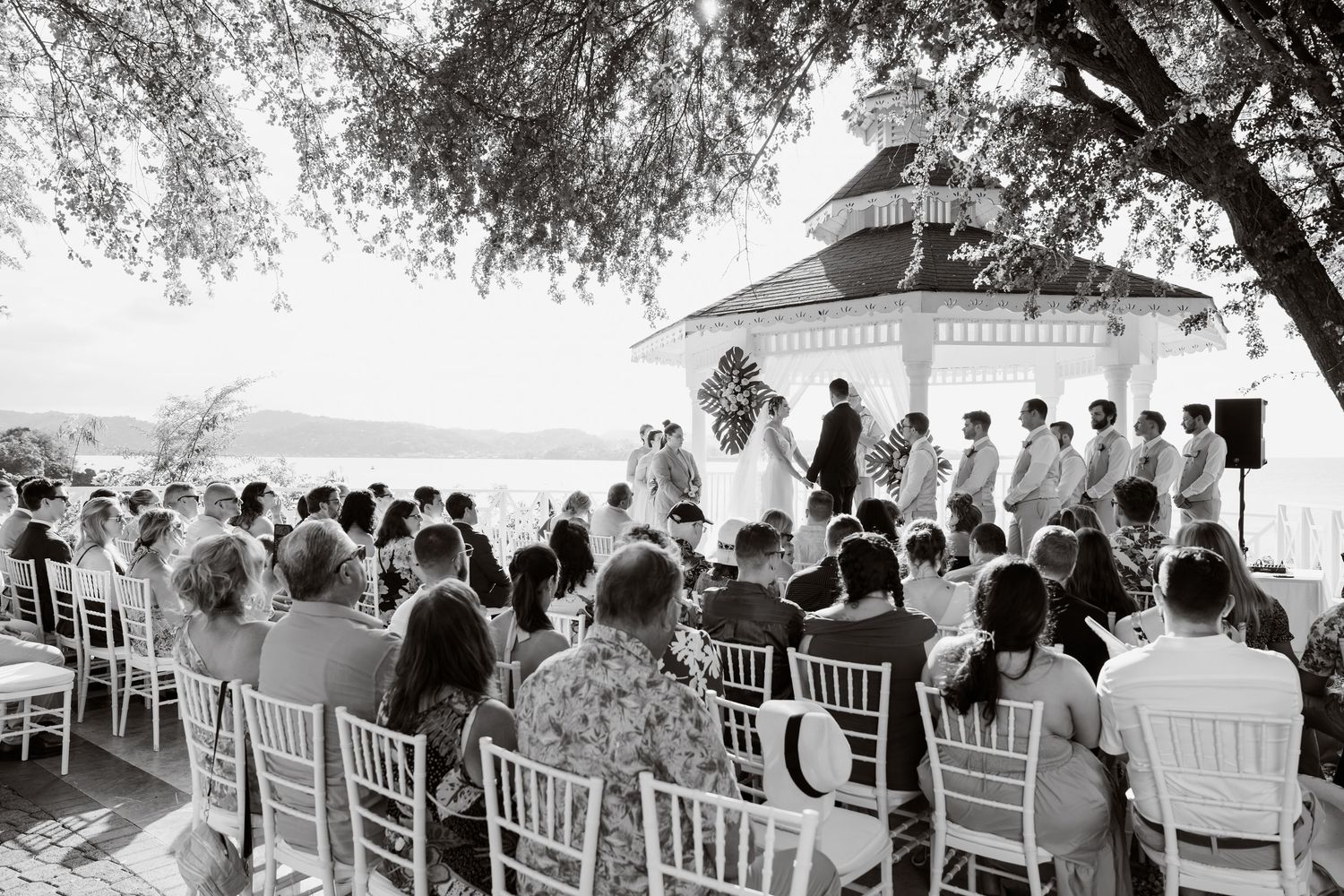 Black and white photo of an outdoor wedding ceremony with white chairs and guests gathered under a gazebo by the water.