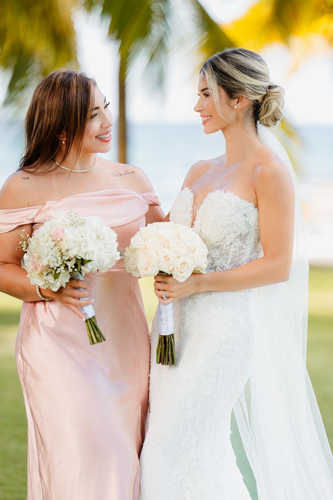 A bridesmaid in pink and bride in white hold matching white bouquets on a sunny lawn.