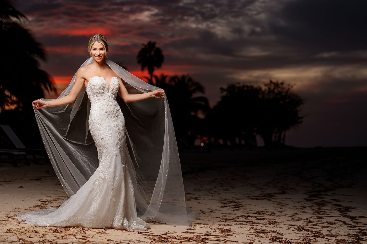 Dramatic beach sunset portrait of bride in lace gown with flowing veil.