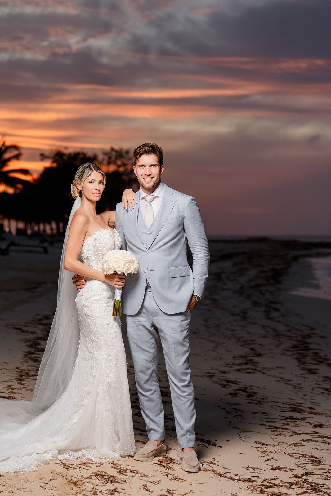 Newlyweds pose for portraits on a beach at sunset with palm trees in silhouette.