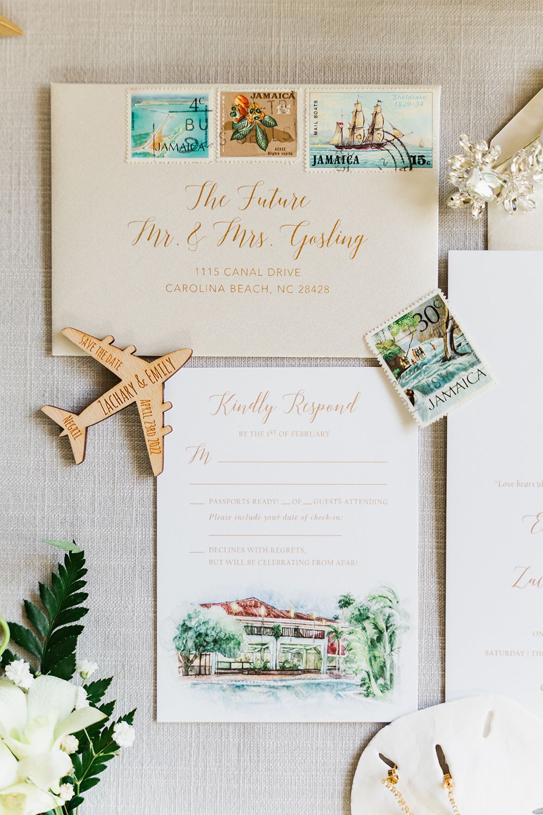 Wedding invitation suite with vintage Jamaica stamps, golden airplane charm, and watercolor venue illustration on light linen background.