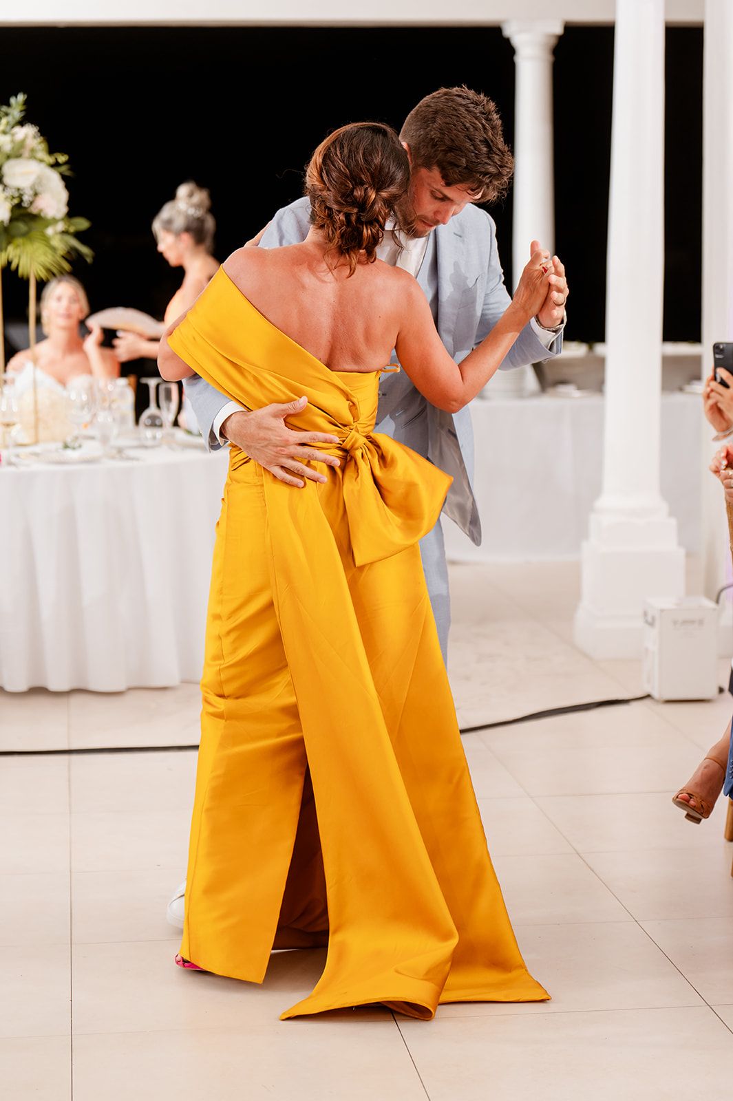 Couple dancing at reception with guest in vibrant yellow jumpsuit.
